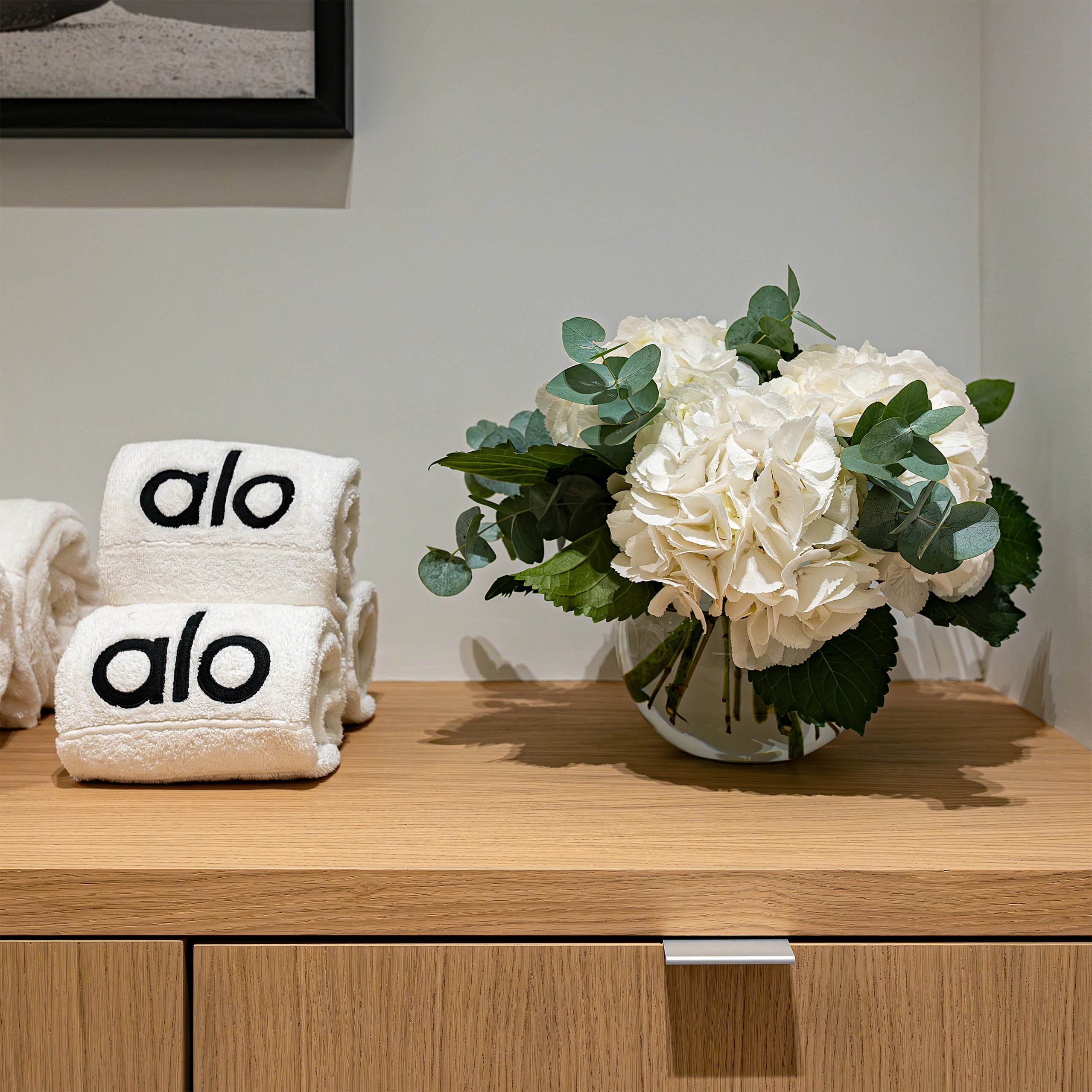 A compact floral arrangement with white hydrangeas and eucalyptus on a wooden counter next to neatly rolled Alo-branded towels at Alo Regent Street Store Grand Opening. The fresh and elegant design, created by Amaranté London Event Florist, enhances the serene and welcoming ambience of the store space.