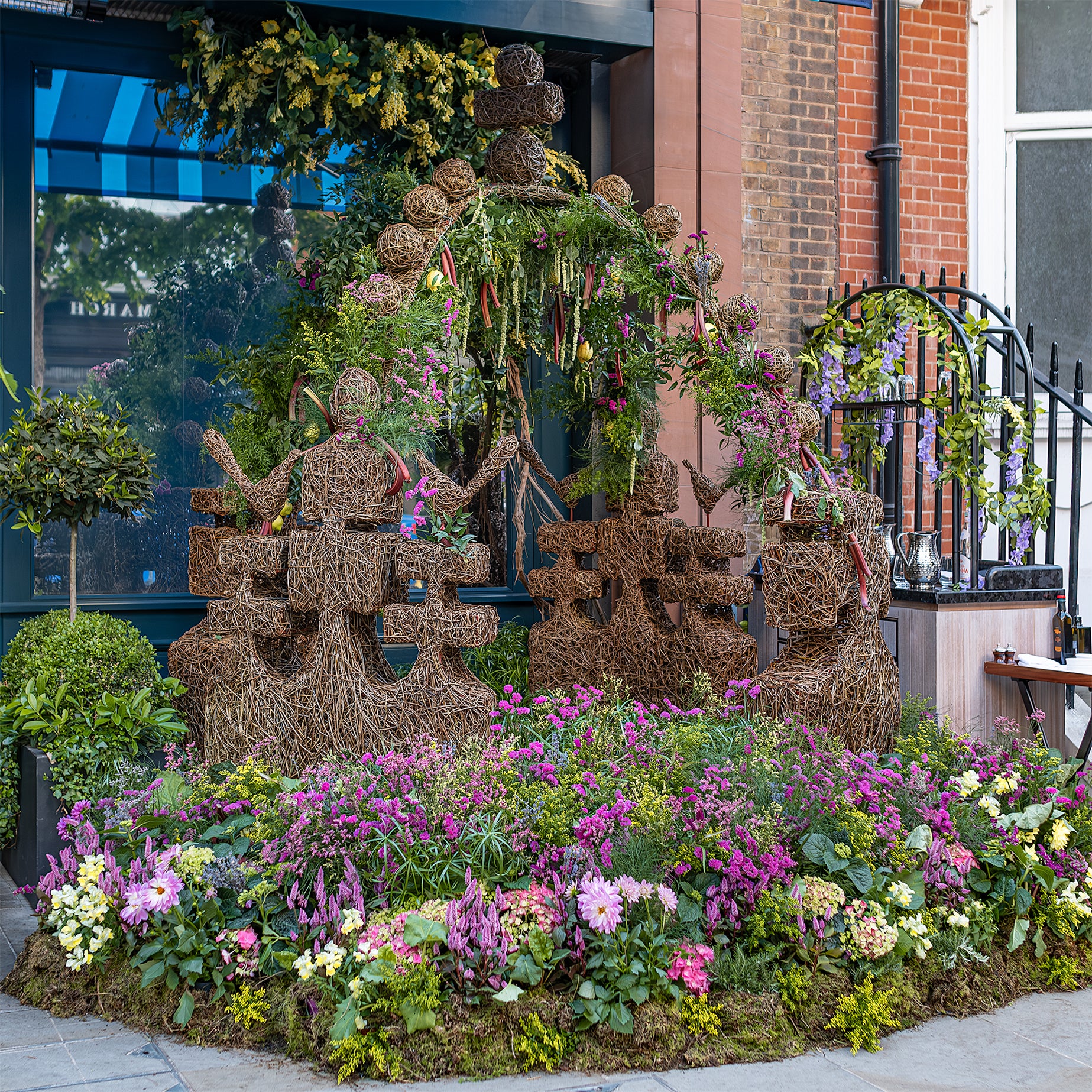 Wicker figures and a floral arch crafted by event florist Amaranté London for Chelsea in Bloom, featuring vibrant pink, yellow, and purple flowers in front of the Azzurra storefront.
