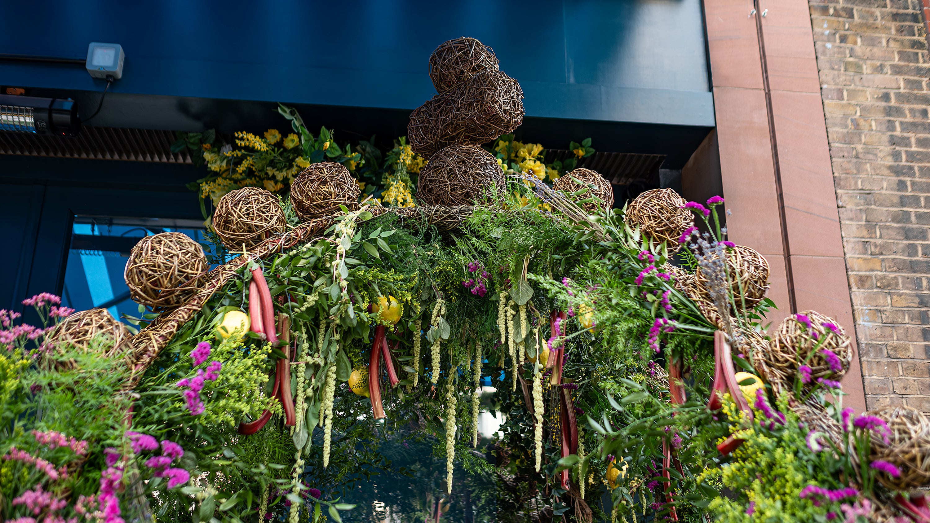 Elegant floral arch designed by Amaranté London for Chelsea in Bloom, decorated with wicker balls, lemons, and a variety of green, yellow, and pink flowers.