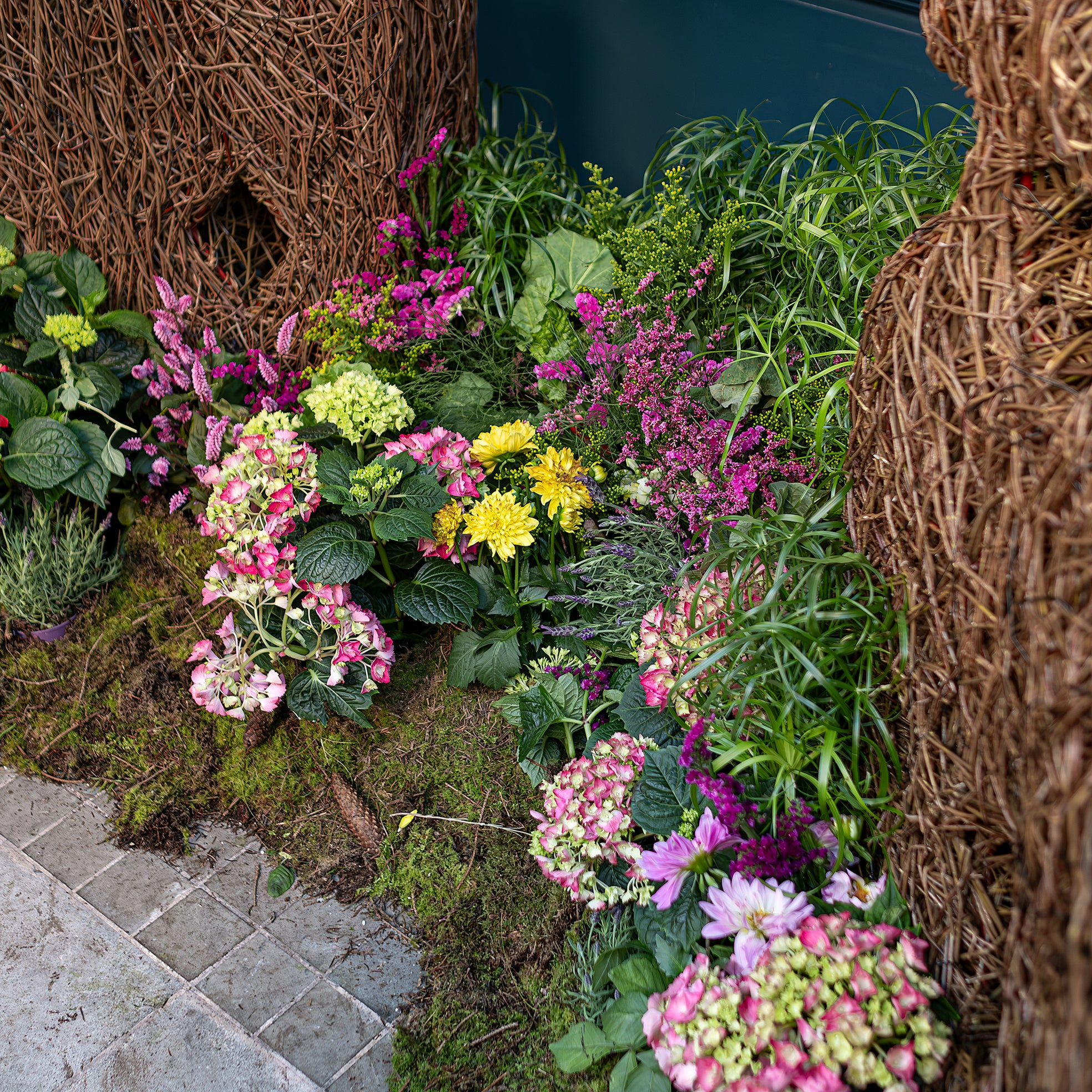 A colourful floral arrangement by Amaranté London for Chelsea in Bloom features pink and yellow flowers, lush green plants, and wicker elements, creating a captivating garden display.