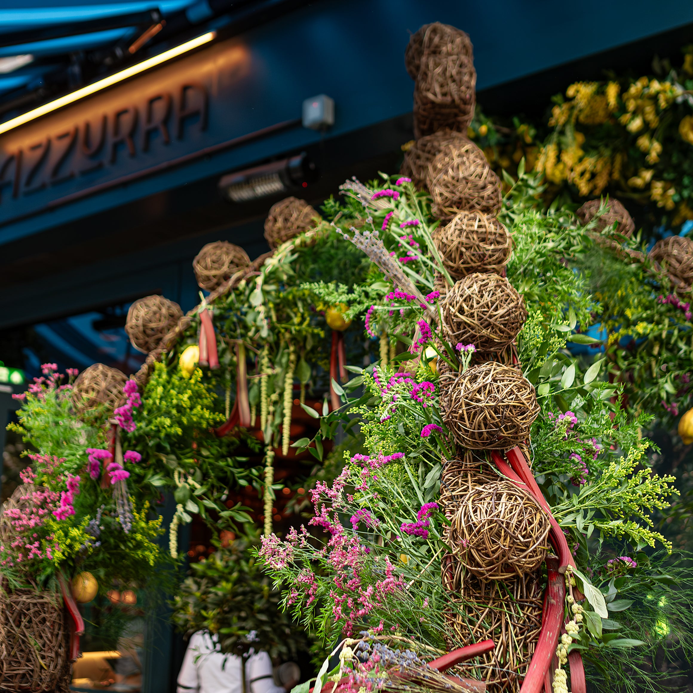 An impressive floral installation by Amaranté London at Chelsea in Bloom showcases a wicker arch adorned with lush greenery, pink and yellow flowers, and lemons in front of the Azzurra storefront.