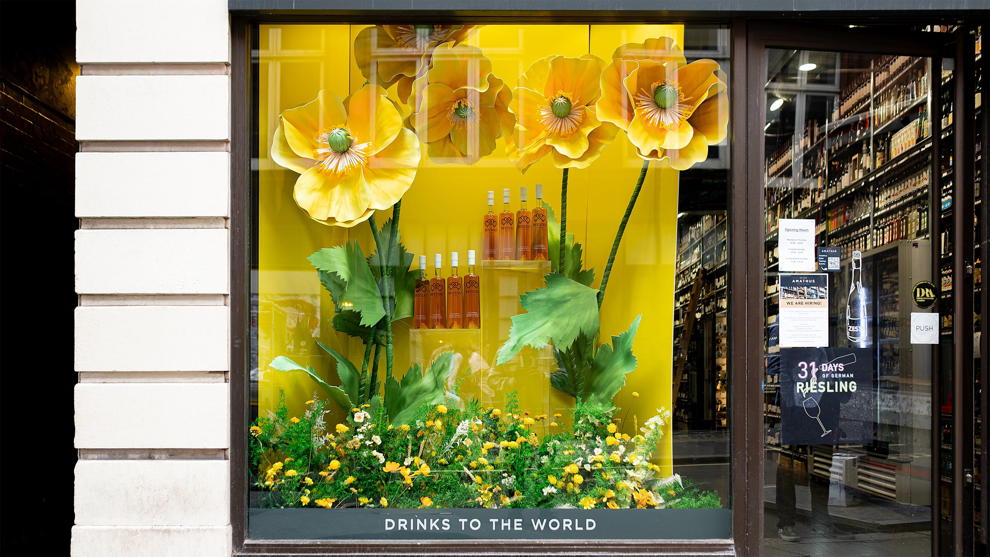 An elegant and eye-catching floral arrangement by Event Florist Amaranté London features large yellow poppies and green plants. Displayed at Amathus, Wardour Street, London, the installation promotes Beesou's aperitif, reflecting the brand's vibrant yellow tones while creating a cohesive visual impact with the product display to attract the attention of people passing by the storefront.