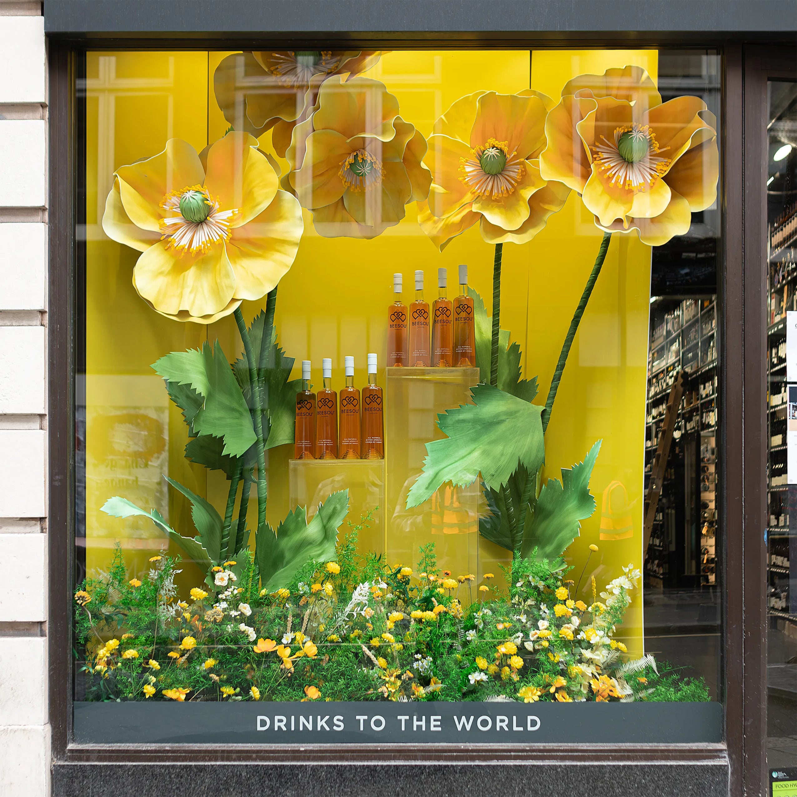 Close-up of Beesou bottles displayed within Amaranté London's bespoke floral window installation at Amathus, Wardour Street, London. The display's yellow poppies and lush greenery highlight the aperitif brand and enhance the visual appeal of the storefront.