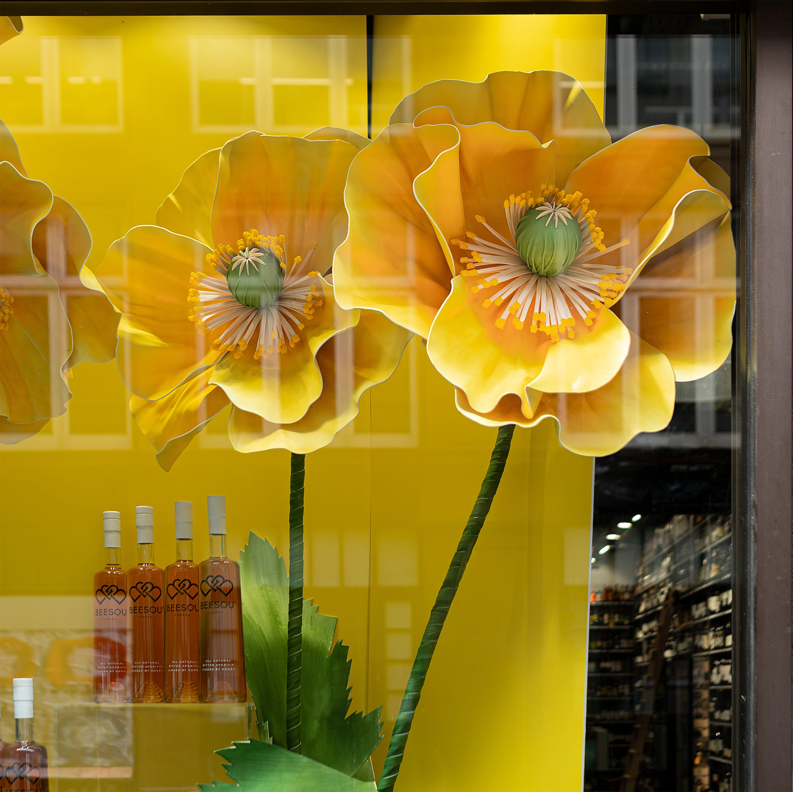 Closer view of the vibrant floral window display featuring large yellow poppy flowers and lush greenery designed by Amaranté London for Beesou's aperitif at Amathus, Wardour Street, London. The installation includes clear acrylic plinths showcasing Beesou bottles, perfectly complementing the brand's colours and enhancing the storefront's visibility.