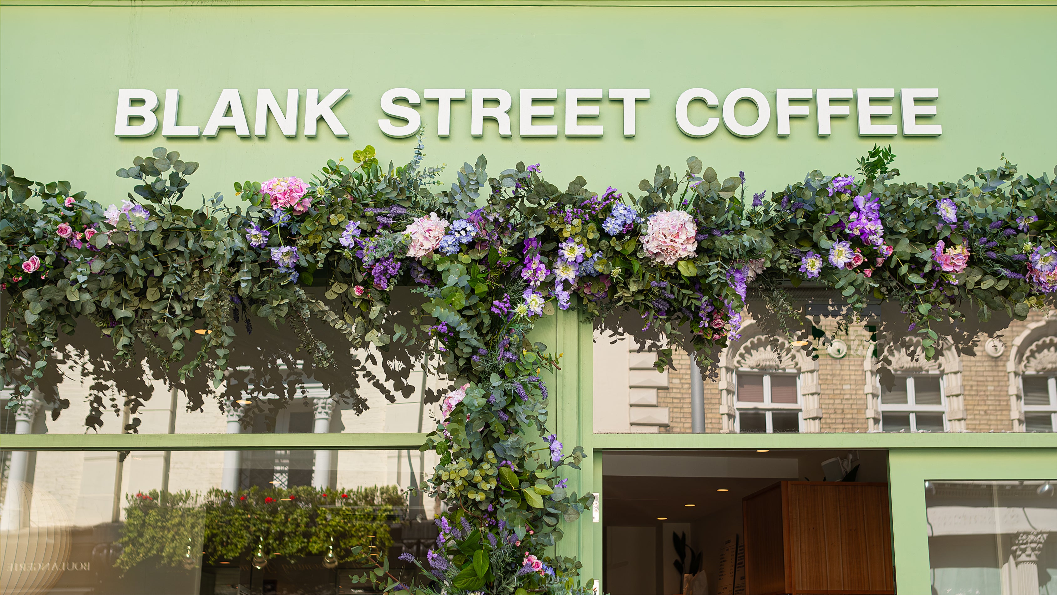 This floral arch, designed by Amaranté London, an event florist in London, was designed for Blank Street Coffee during Chelsea in Bloom. It showcases an elegant blend of pink roses, purple hydrangeas, and green foliage. The floral arrangement frames the café's entrance, creating a visually captivating, welcoming experience.