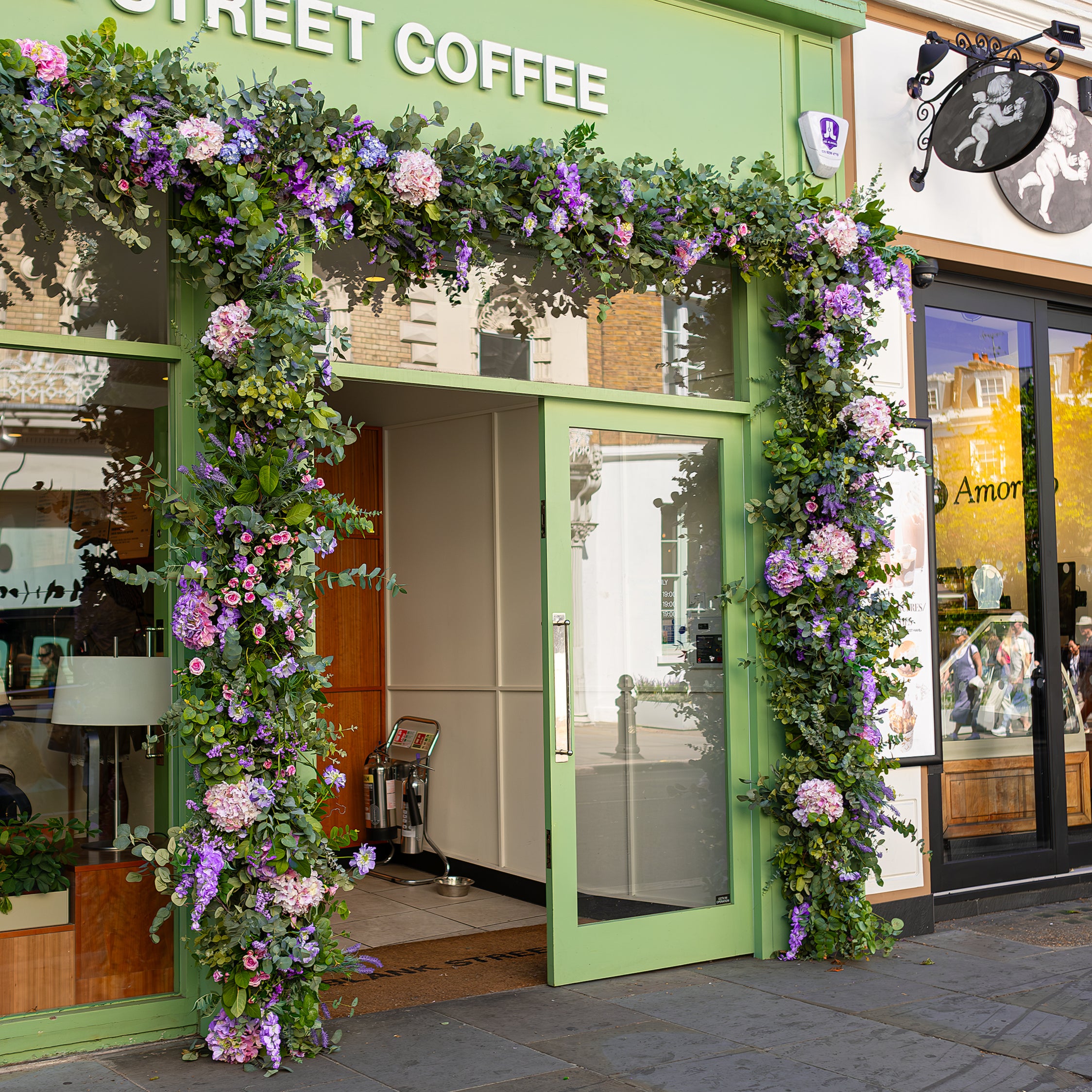 Image of Amaranté London's floral installation at Blank Street Coffee for Chelsea in Bloom, featuring a mix of lush green eucalyptus, pink roses, and purple flowers. The vibrant arrangement enhances the café's storefront, making it a focal point of natural beauty on King's Road.