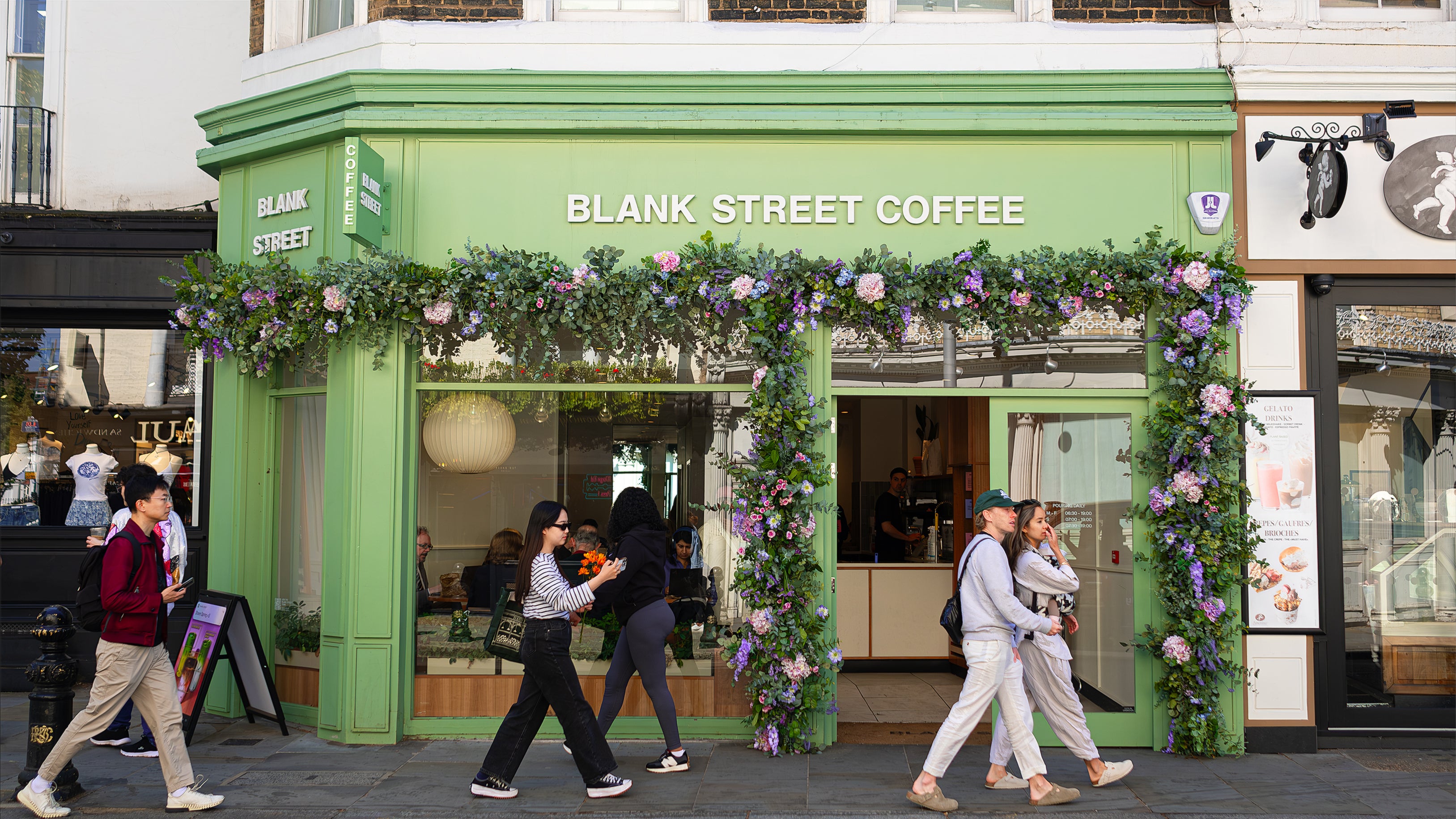 Wide view of Blank Street Coffee's storefront adorned with Amaranté London's floral arrangement for Chelsea in Bloom. The installation features an archway of lush green foliage, pink and purple flowers, enhancing the café's exterior and creating a striking visual impact.