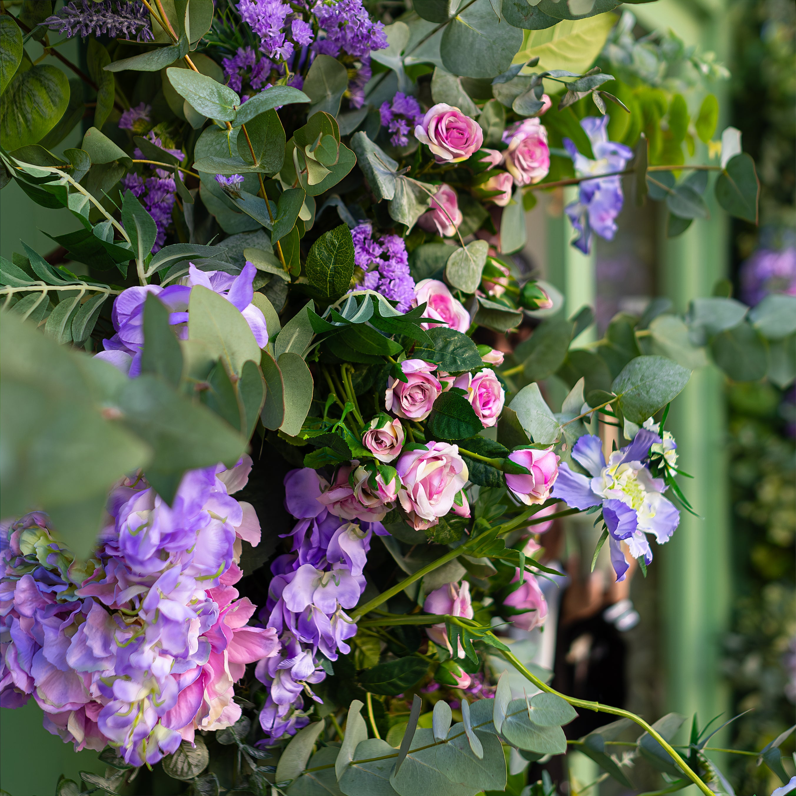 This is a close-up of the vibrant floral arch arrangement created by event florist Amaranté London for Blank Street Coffee's storefront during Chelsea in Bloom. It features lush green foliage with pink roses, purple hydrangeas, and delicate lavender sprigs, enhancing the natural beauty of the store's exterior.