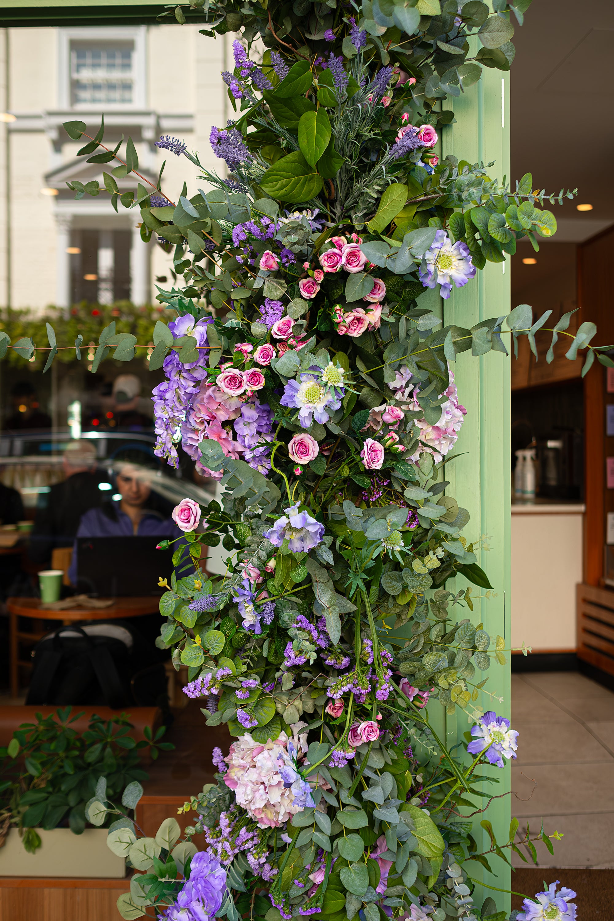 Image of a detailed view of Amaranté London's floral installation for Blank Street Coffee at Chelsea in Bloom. The arrangement showcases a cascade of green eucalyptus leaves, pink roses, and purple flowers, creating a lush, inviting entrance that draws attention to the café.