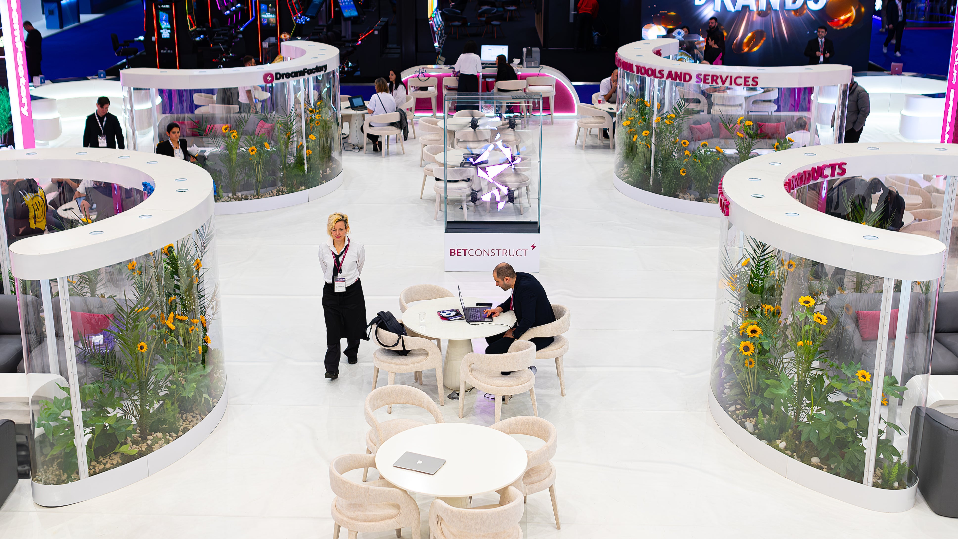In the image, circular floral installations serve as decoration and partition within the corporate event space. These arrangements prominently feature yellow sunflowers complemented by a variety of green foliage. The flowers are encased in clear, cylindrical displays, interspersed among seating areas and workstations, providing a natural and vibrant contrast to the event's predominantly white and modern setting at the ICE London Exhibition.