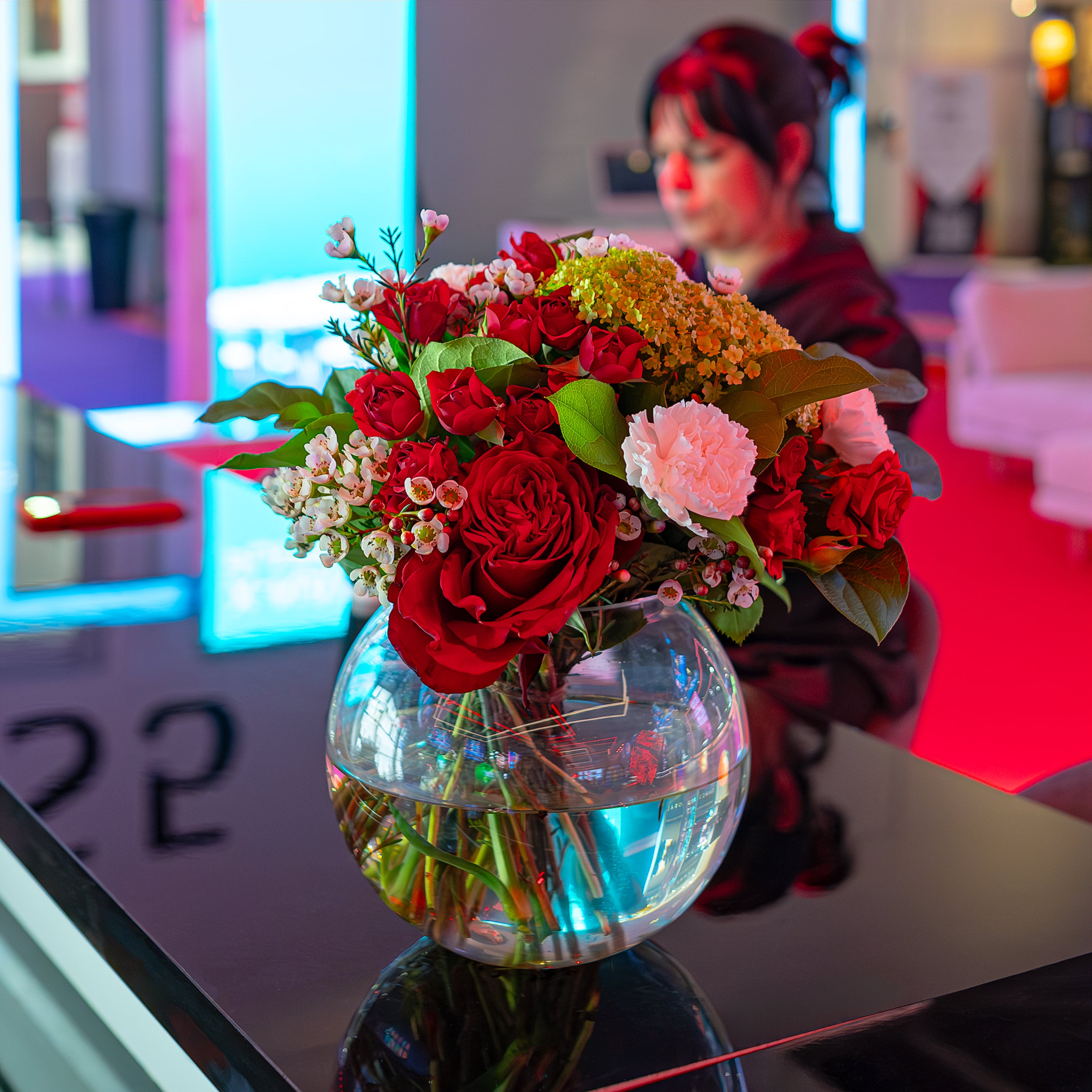 A stunning bespoke luxury bouquet of deep red roses and delicate pink blooms is presented in a glass vase against the vibrant backdrop in an area of the ICE London Exhibition