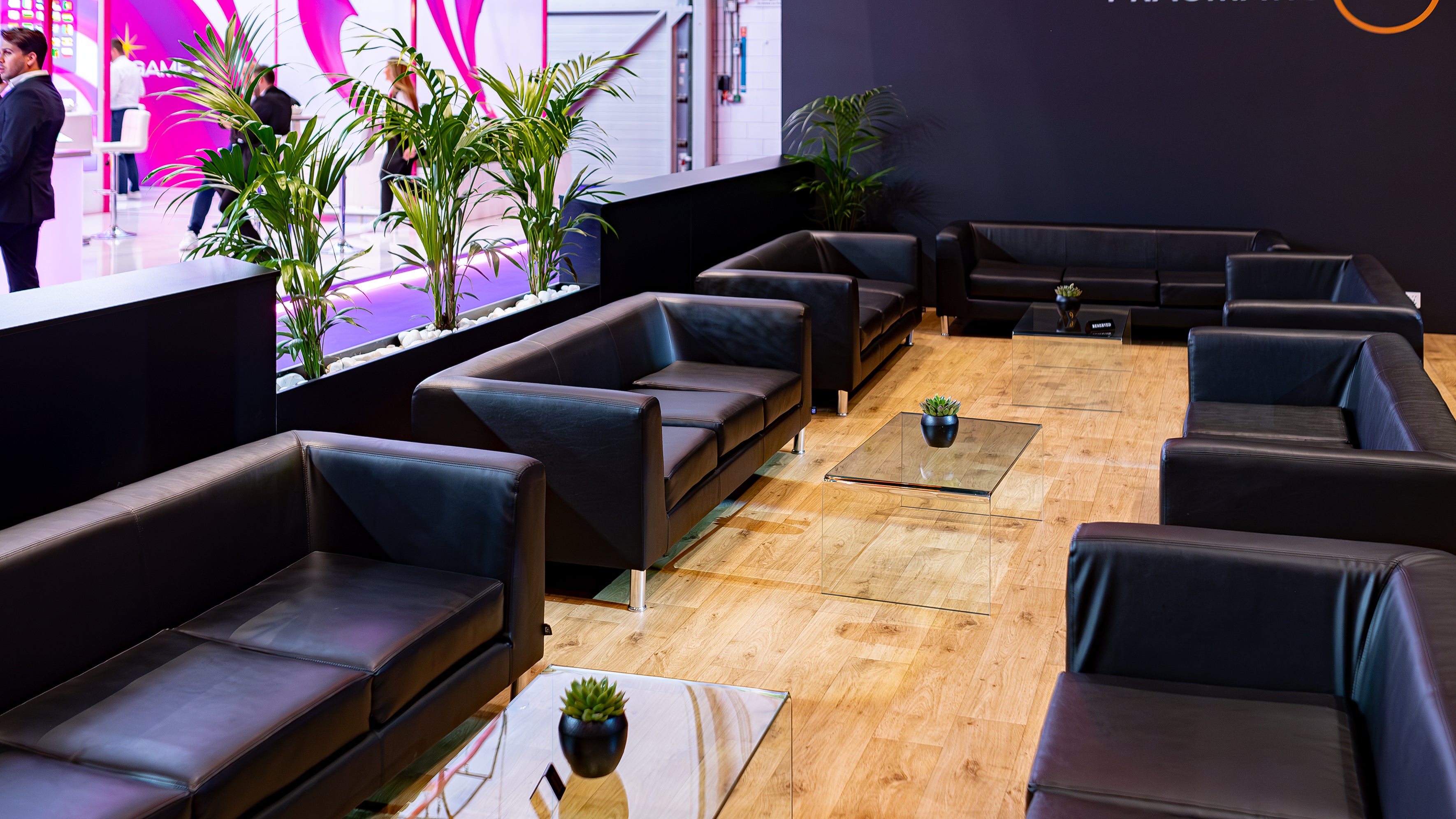A contemporary lounge setup at ICE London Exhibition was designed by event florist Amaranté London, featuring sleek black leather sofas and glass tables, complemented by green potted plants, creating a stylish and welcoming discussion area.