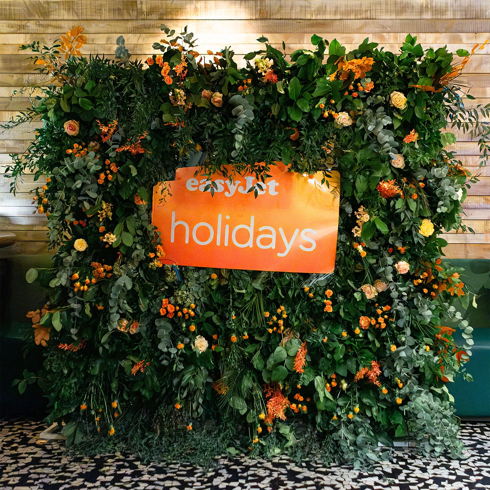 A flower wall at the venue entrance to welcome guests to the EasyJet annual partner event featuring vibrant orange and cream roses surrounded by greenery - Floral Design By AmarantÃ© London.