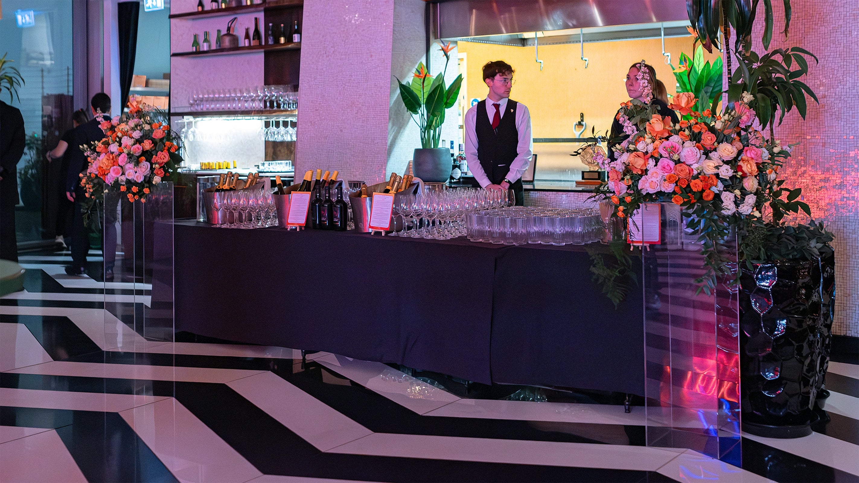 Floral arrangements with vibrant orange and blush roses, pink lisianthus, and eucalyptus greenery, complemented by polished green-leafed plants in ceramic planters on the warm wooden bar near neatly arranged champagne flutes to enhance the luxurious and contemporary setting for EasyJetâ€™s annual partner event - Floral Design by AmarantÃ© London"