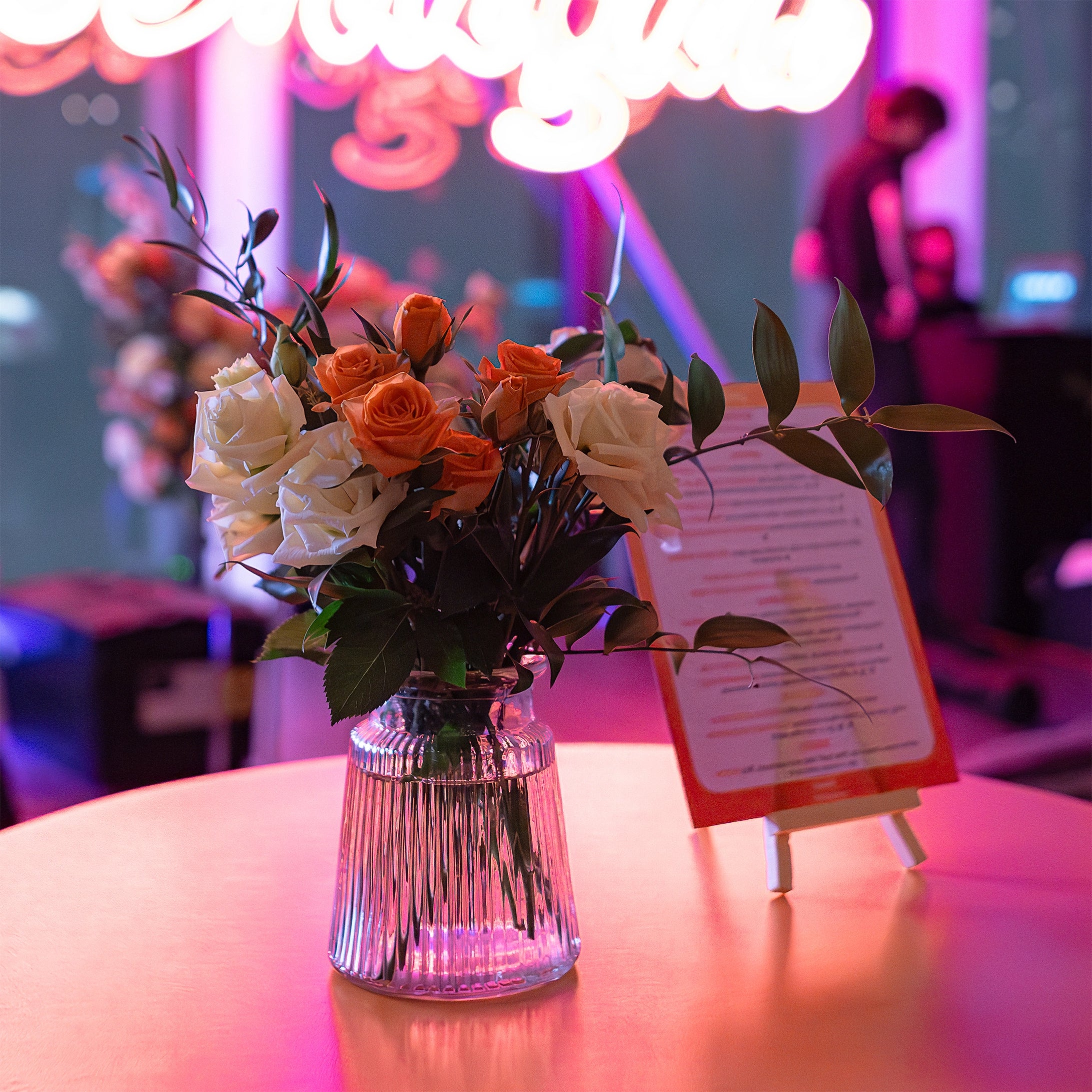 A flower table arrangement with vibrant orange and cream roses seamlessly combines with lush greenery, creating a captivating centrepiece for the EasyJet annual partner event in London - Floral Design by AmarantÃ© London