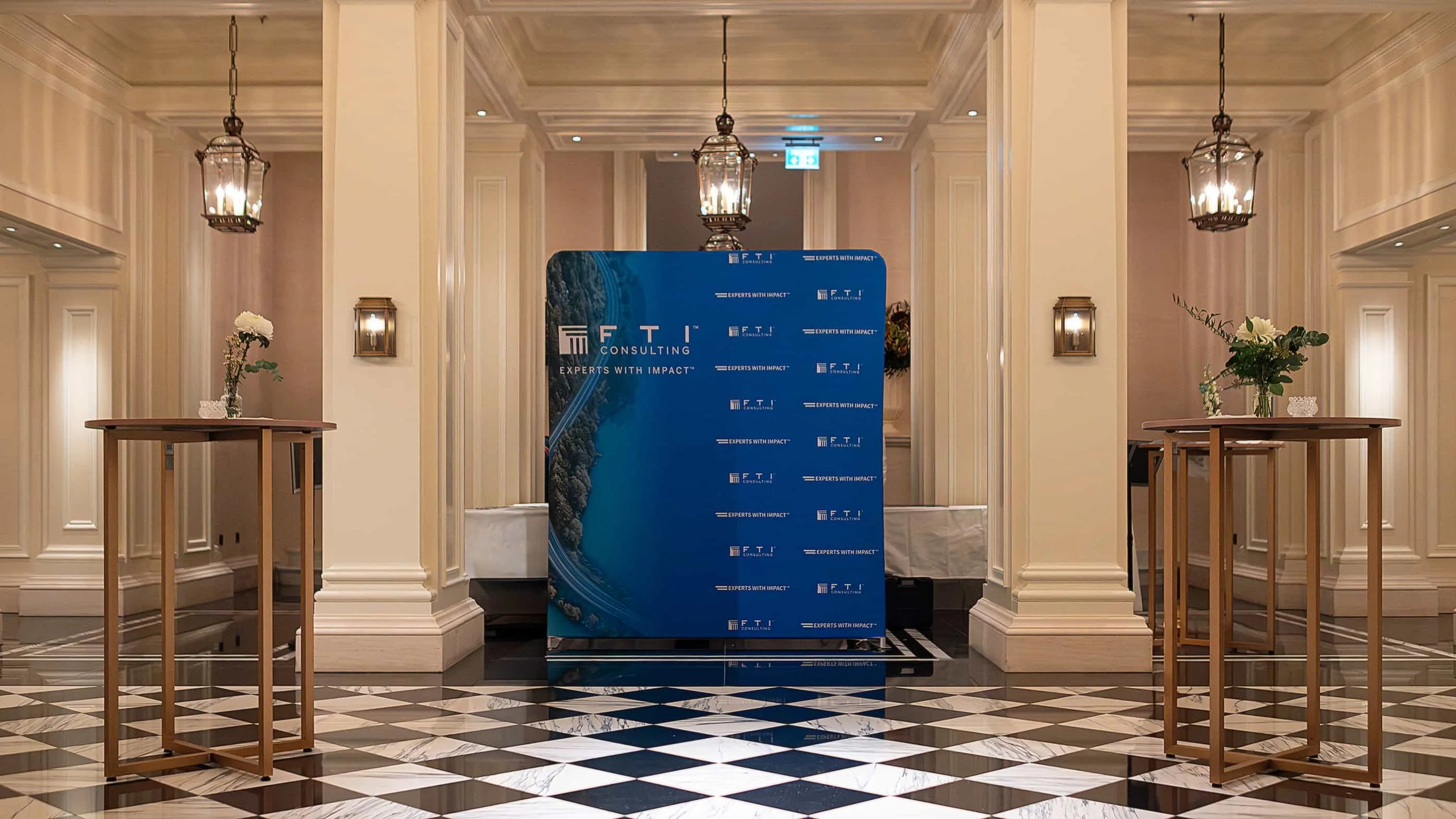 A welcoming entrance to the FTI Consulting corporate event, featuring a branded backdrop for guest photos, flanked by two high tables each adorned with an elegant vase of flowers by Amaranté London - Event Florist in London