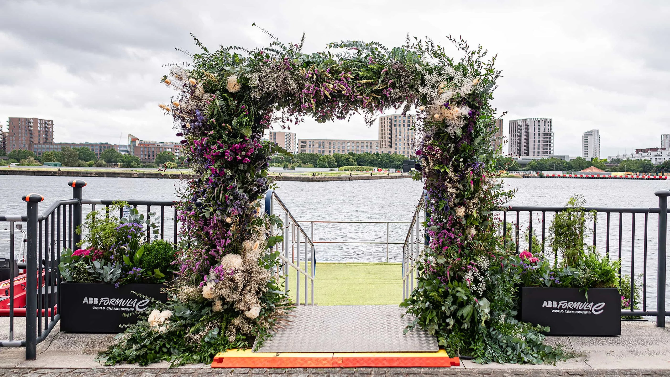 The majestic, floral archway at the ABB Formula E World Championship entrance, adorned with lush, vibrant flowers in purples, whites, and greens, overlooks the serene waters of the London docks, enhancing the event's grandeur and sustainability ethos - Amaranté London.