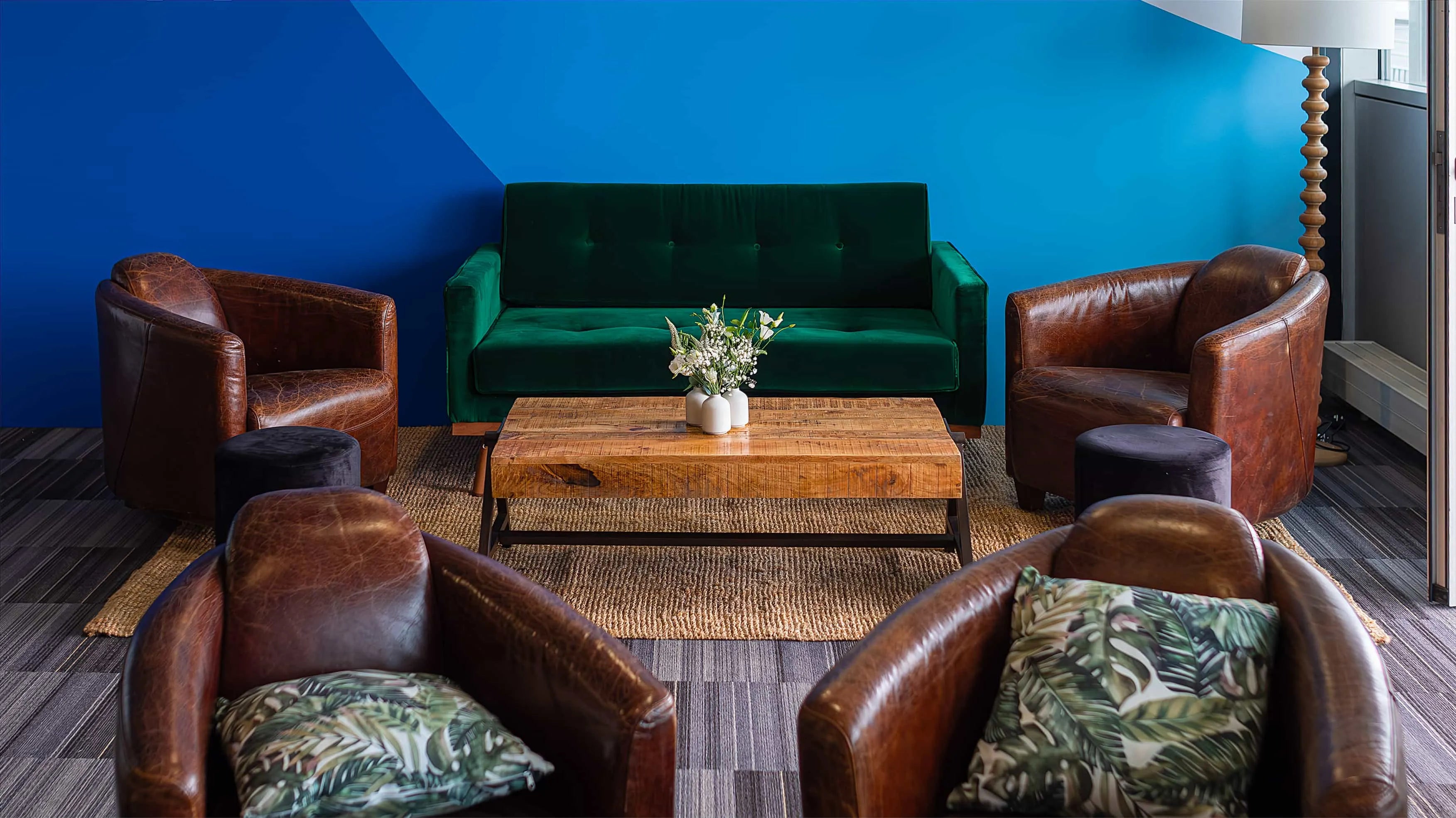 Chic and cosy lounge area featuring a bold emerald green sofa and vintage brown leather chairs, complemented by a simple yet stylish white floral arrangement on a rustic coffee table set against a vibrant blue wall - Amaranté London.