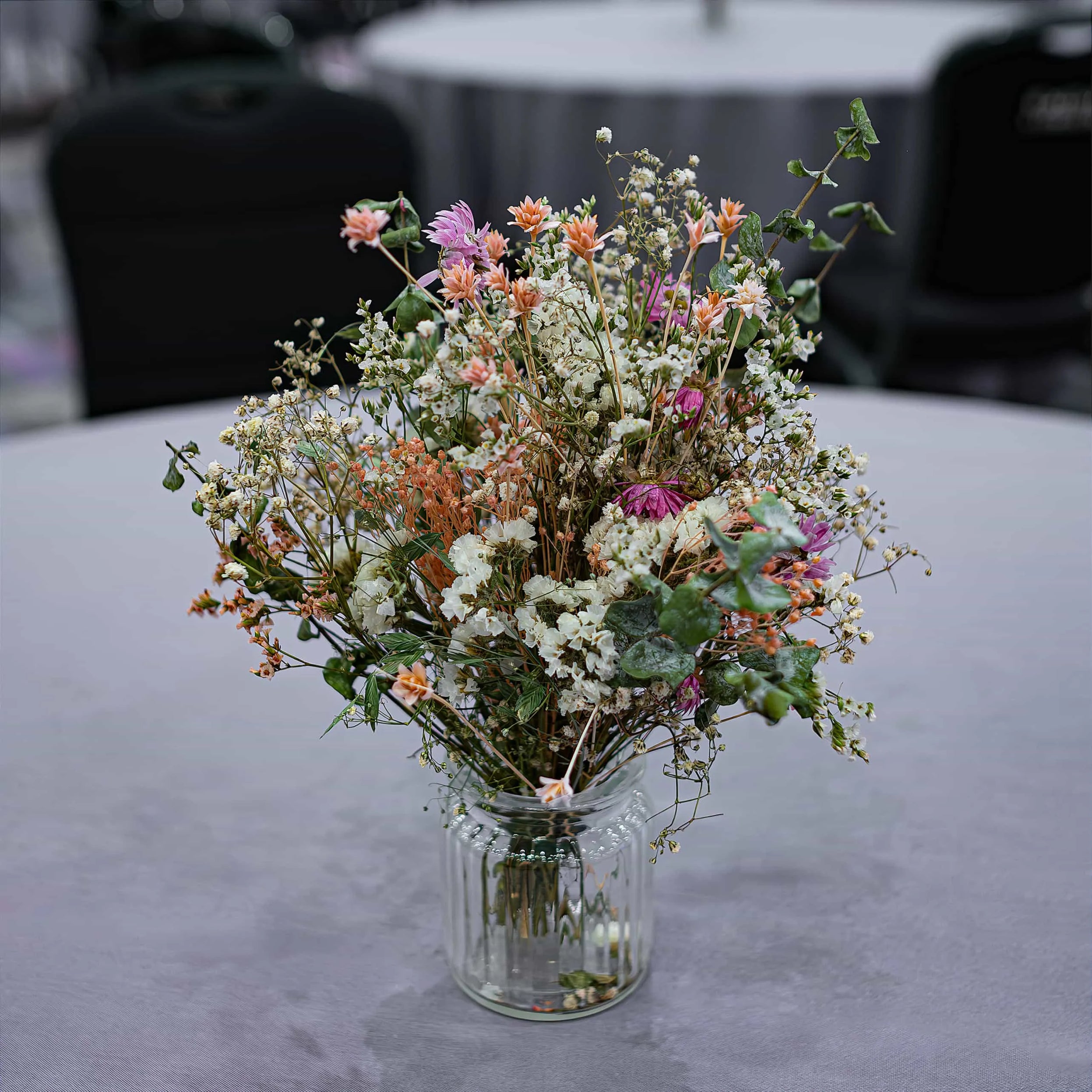 Rustic wildflower arrangement in a clear jar by Amaranté London at the Formula E event, combining orange, pink, and white blooms with green accents on a grey clothed table, enhancing the naturalistic setting.