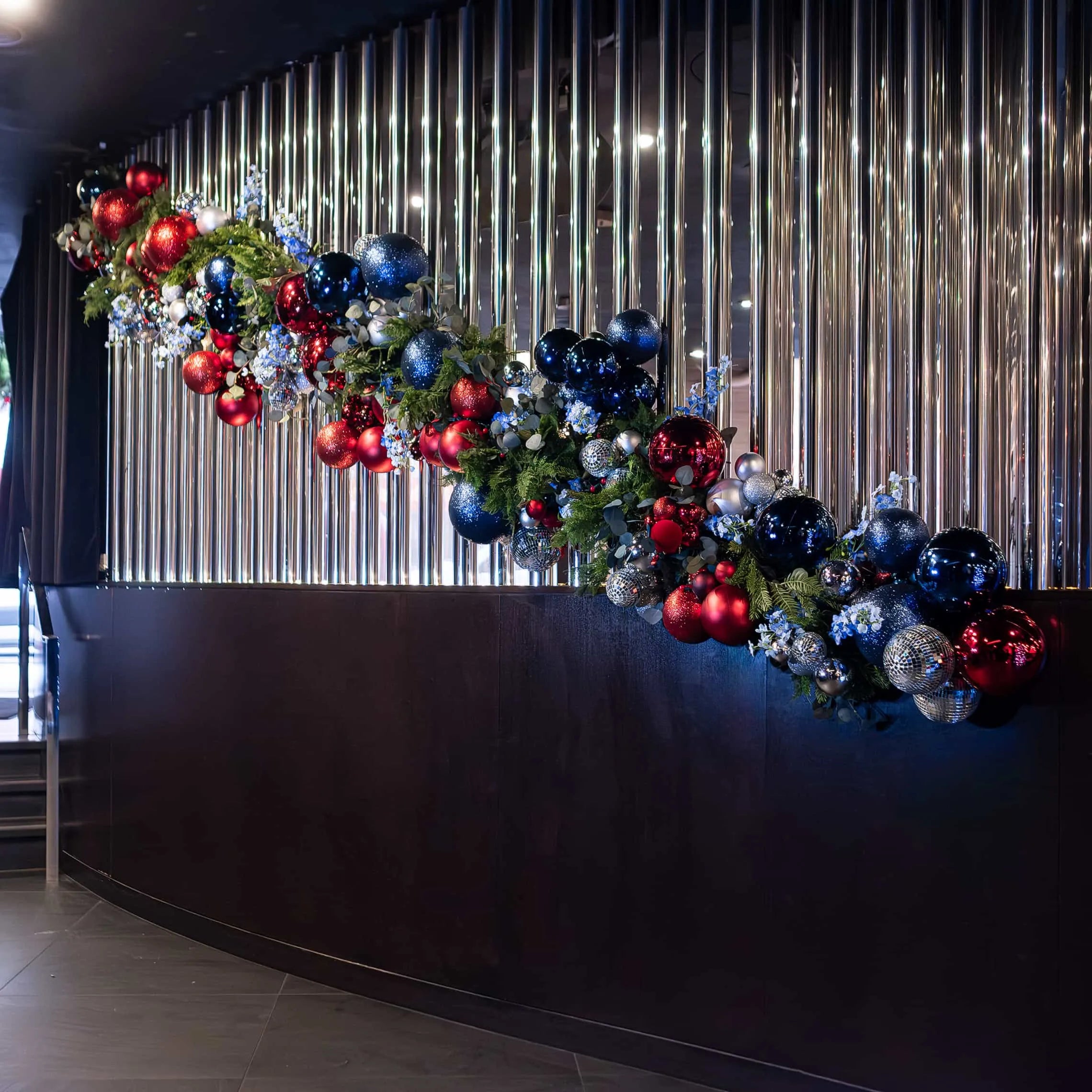 Festive Christmas garland elegantly draped over the reception desk at STK Steakhouse, with lush greenery against a modern backdrop of vertical metallic stripes, red and blue baubles, and silver decorations.