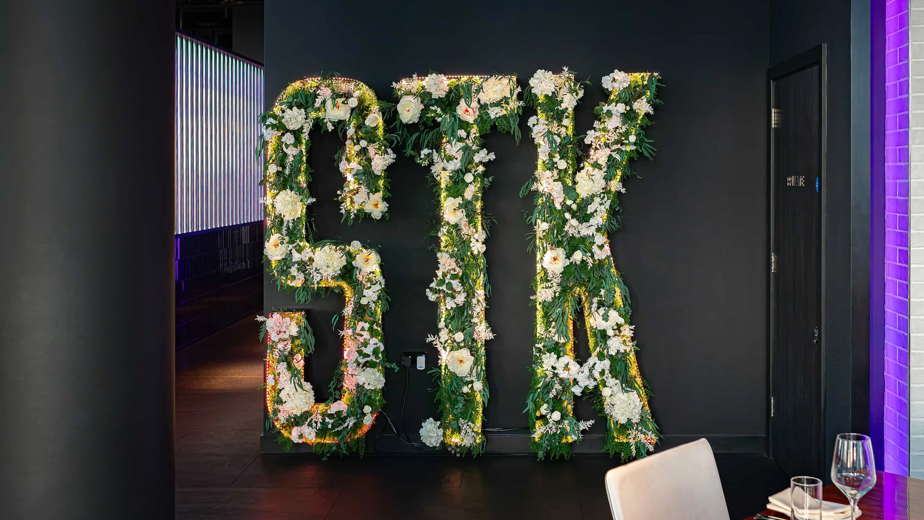 The letters 'STK', the Brand of this famous Steakhouse in London, are artistically fashioned from a vibrant display of white flowers and lush greenery, illuminated by subtle fairy lights, creating a striking Christmas floral arrangement against the sleek, dark walls of STK Steakhouse entrance.