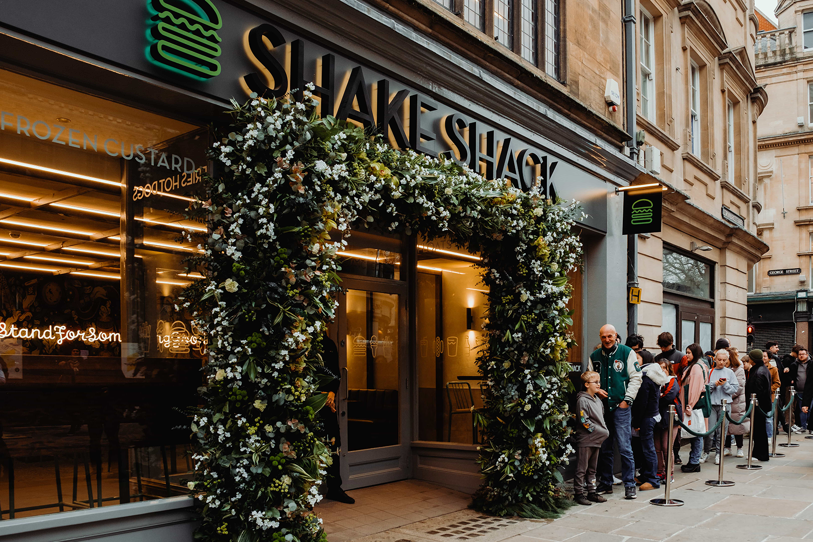 This is an exterior view of the Shake Shack new store opening in Manchester. The image features the store entrance adorned with a custom floral arch ddesigned and created by event florist Amaranté London, known for their bespoke floral design services throughout the UK. This large floral arch has deep green foliage and white floral accents, creating a stunning visual impact for the grand opening.