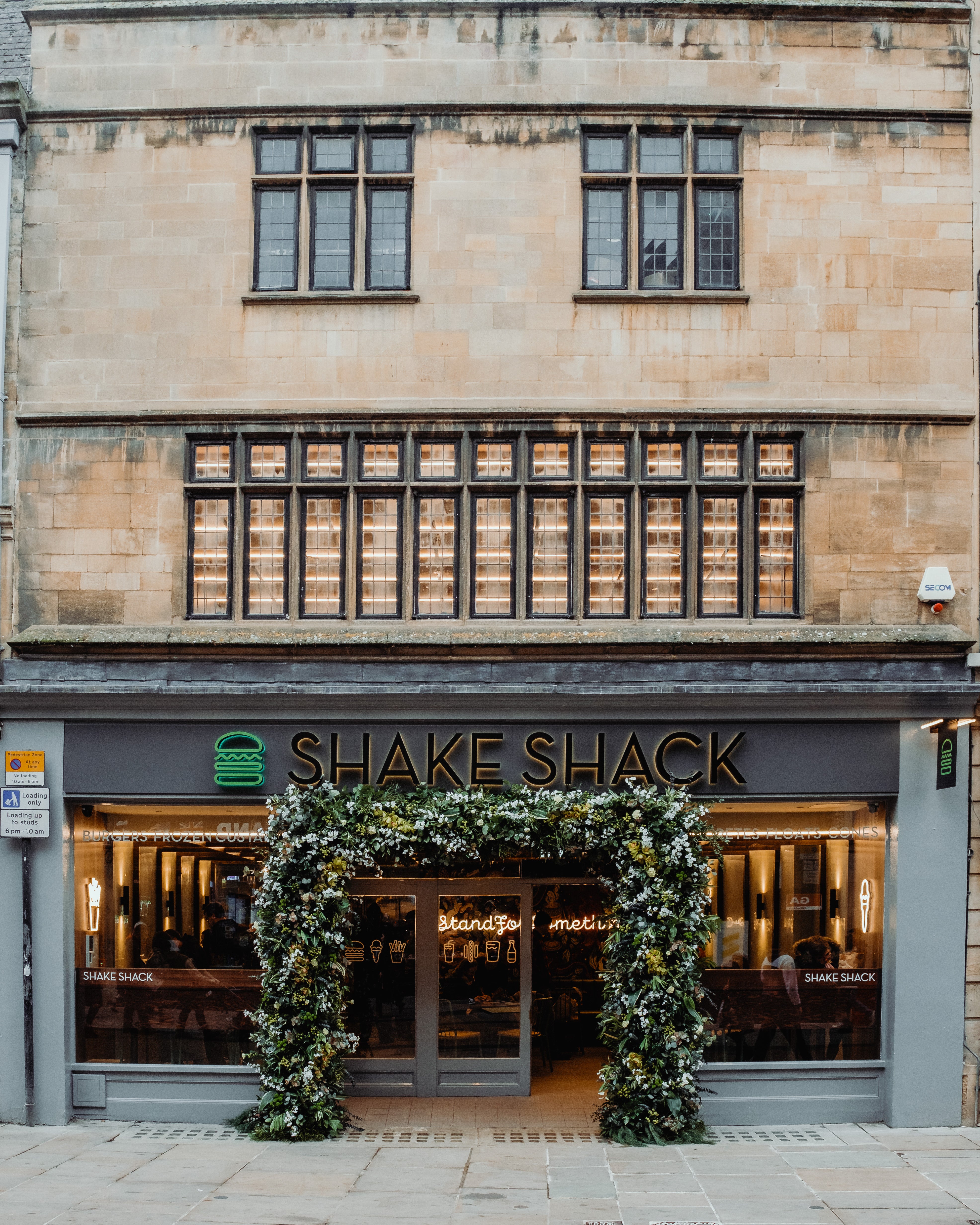 This is an exterior view of the Shake Shack new store opening in Manchester. The image features the store entrance adorned with a custom floral arch ddesigned and created by event florist Amaranté London, known for their bespoke floral design services throughout the UK. This large floral arch has deep green foliage and white floral accents, creating a stunning visual impact for the grand opening.