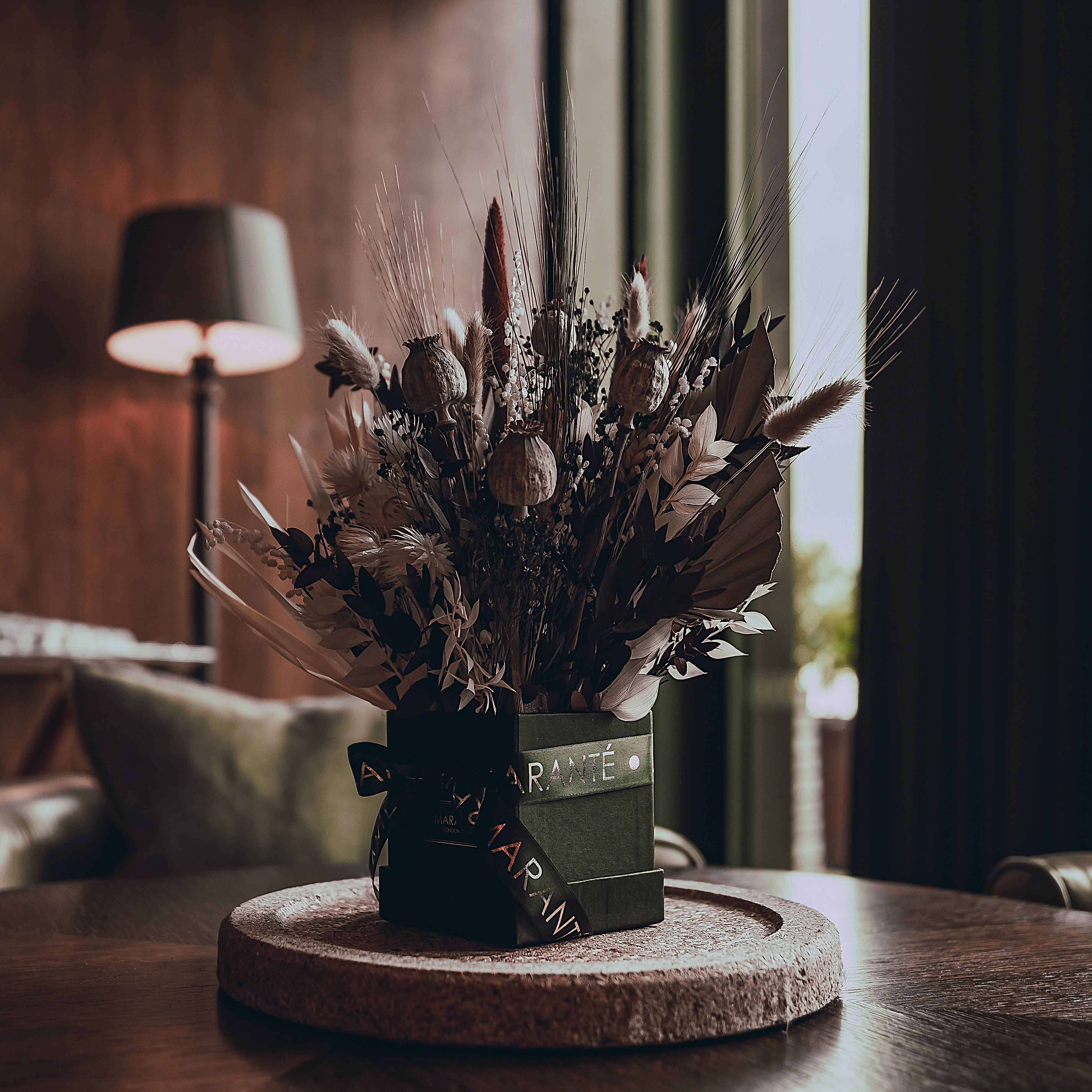 An Amaranté bespoke floral arrangement for events stands majestically on a circular cork base, featuring an array of dried blooms in earthy tones, perfectly complementing the moody atmosphere of a luxurious space at SOHO HOUSE.