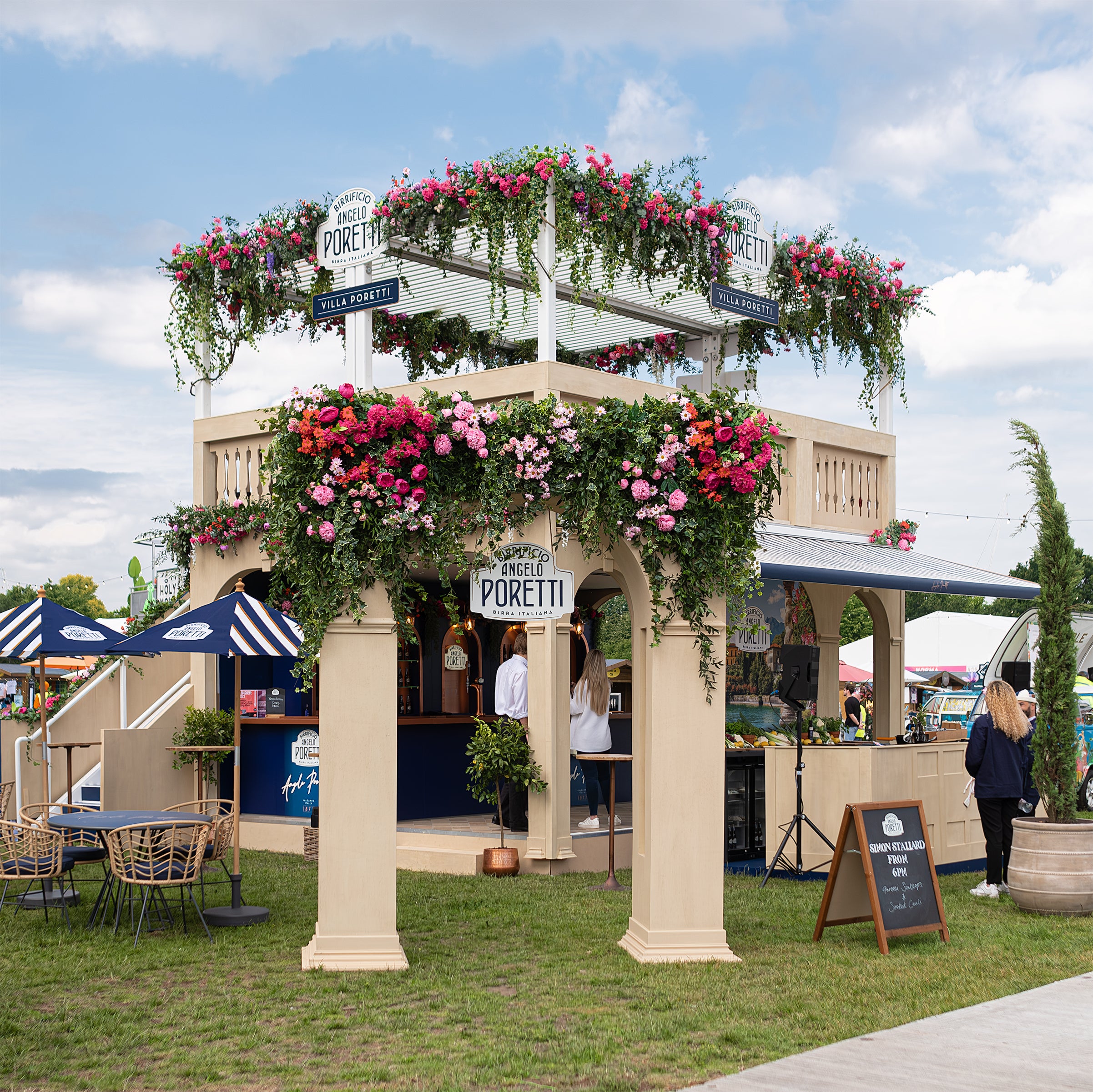 Angelo Poretti beer stand at Regent’s Park Taste Festival decorated by Event Florist Amaranté London. The archway is adorned with vibrant pink, red, and orange flowers and lush green foliage. Blue awnings and wicker chairs enhance the Mediterranean ambiance. An exquisite example of Amaranté London’s event floristry.