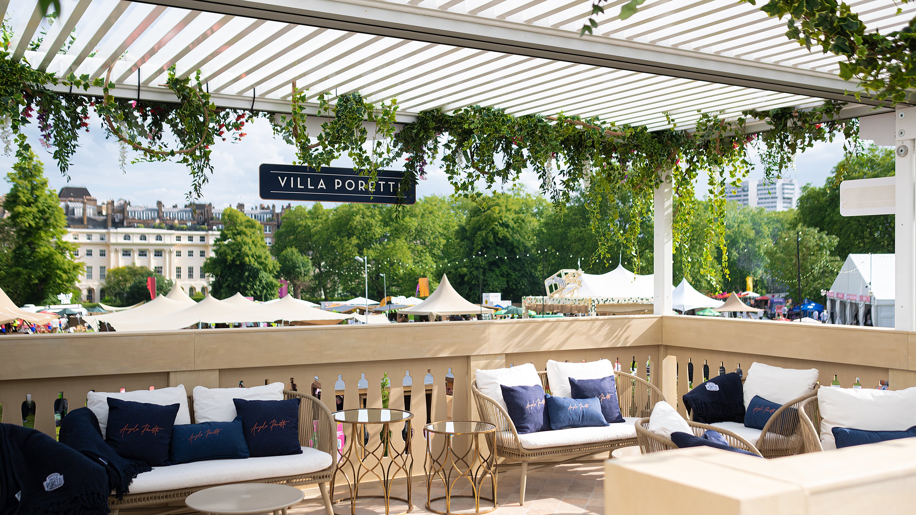 Image of the upper terrace of Angelo Poretti stand at Regent’s Park Taste Festival Event, featuring a luxurious seating area with blue and white cushions. Hanging ivy and floral arrangements, all designed, created and installed by Amaranté London. The bespoke floral arrangements add to the Mediterranean ambience.