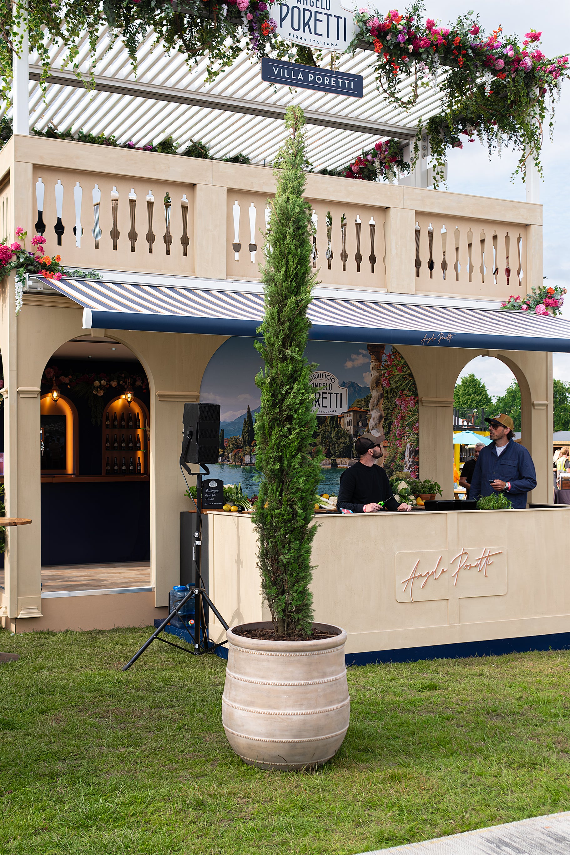 Exterior view of the Angelo Poretti stand at Regent’s Park Taste Festival, featuring a tall potted cypress tree in a beige planter by Amaranté London. The floral decor includes vibrant flowers in pink, orange, and white, creating a Mediterranean feel. An example of Amaranté London’s plant hire and event floristry services.