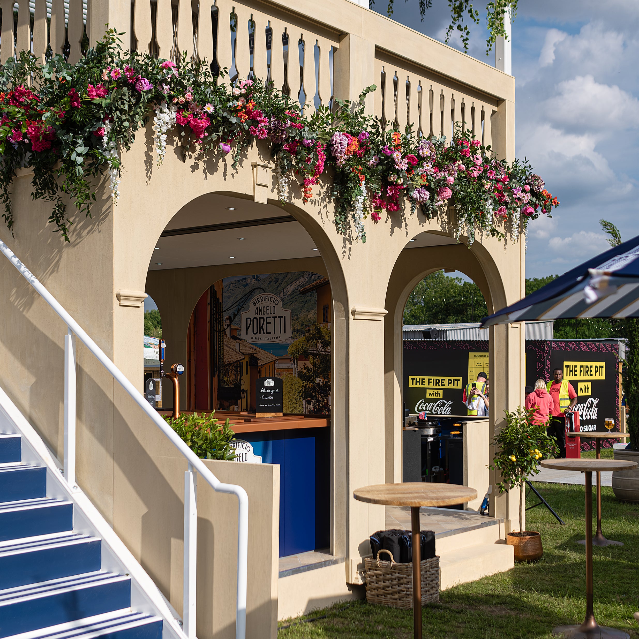 Angelo Poretti beer stand at Regent’s Park Taste Festival featuring floral decor for events by florist Amaranté London. The floral decorations for this stand include arches adorned with vibrant fresh flowers and green foliage, wicker seating, and blue awnings.