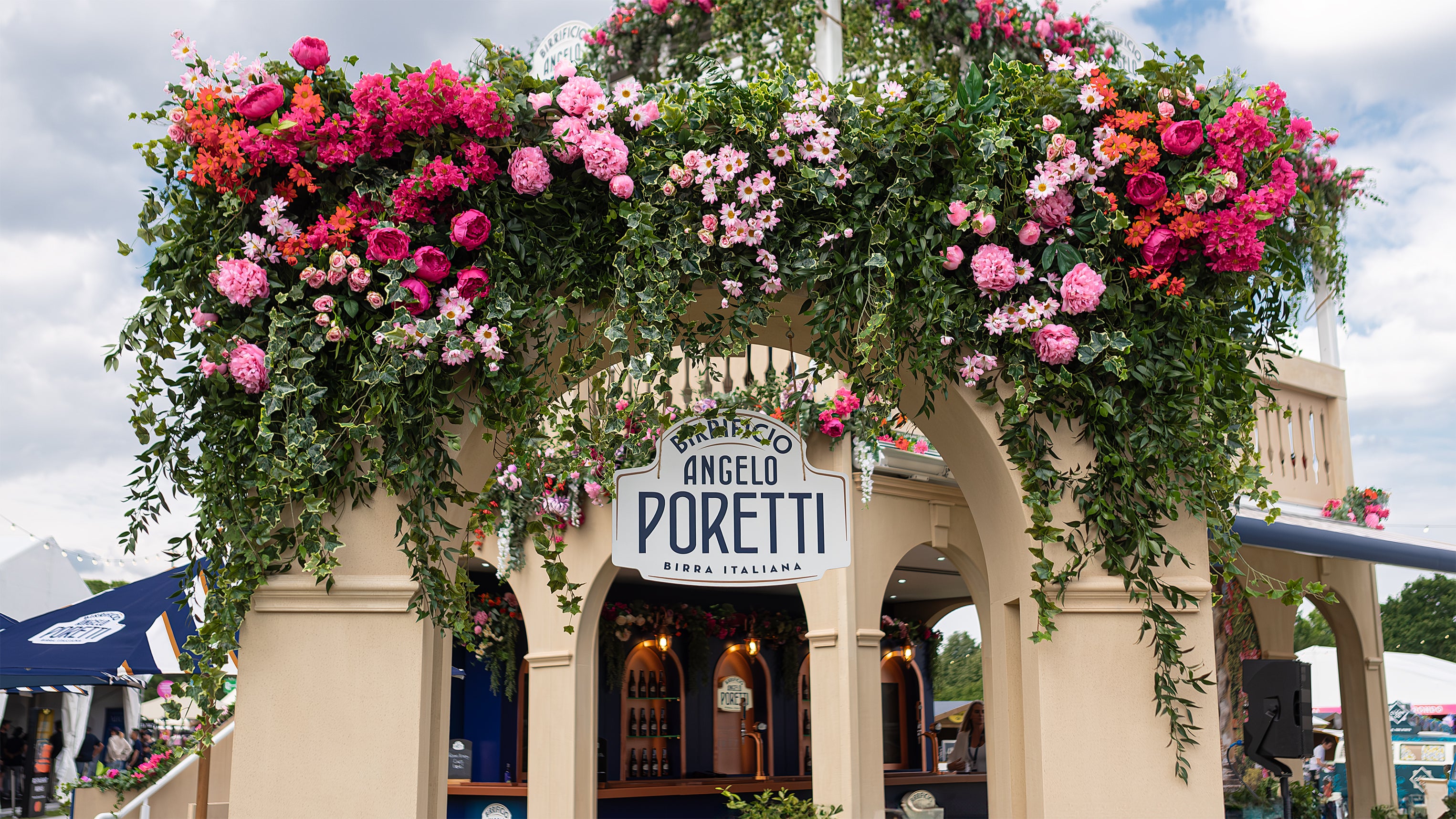 This is a close-up view of the Angelo Poretti sign at Regent’s Park Taste Festival. Above the archway, lush green ivy and vibrant flowers in shades of pink, red, and orange create a welcoming atmosphere—bespoke floral arrangements by Event Florist Amaranté London.