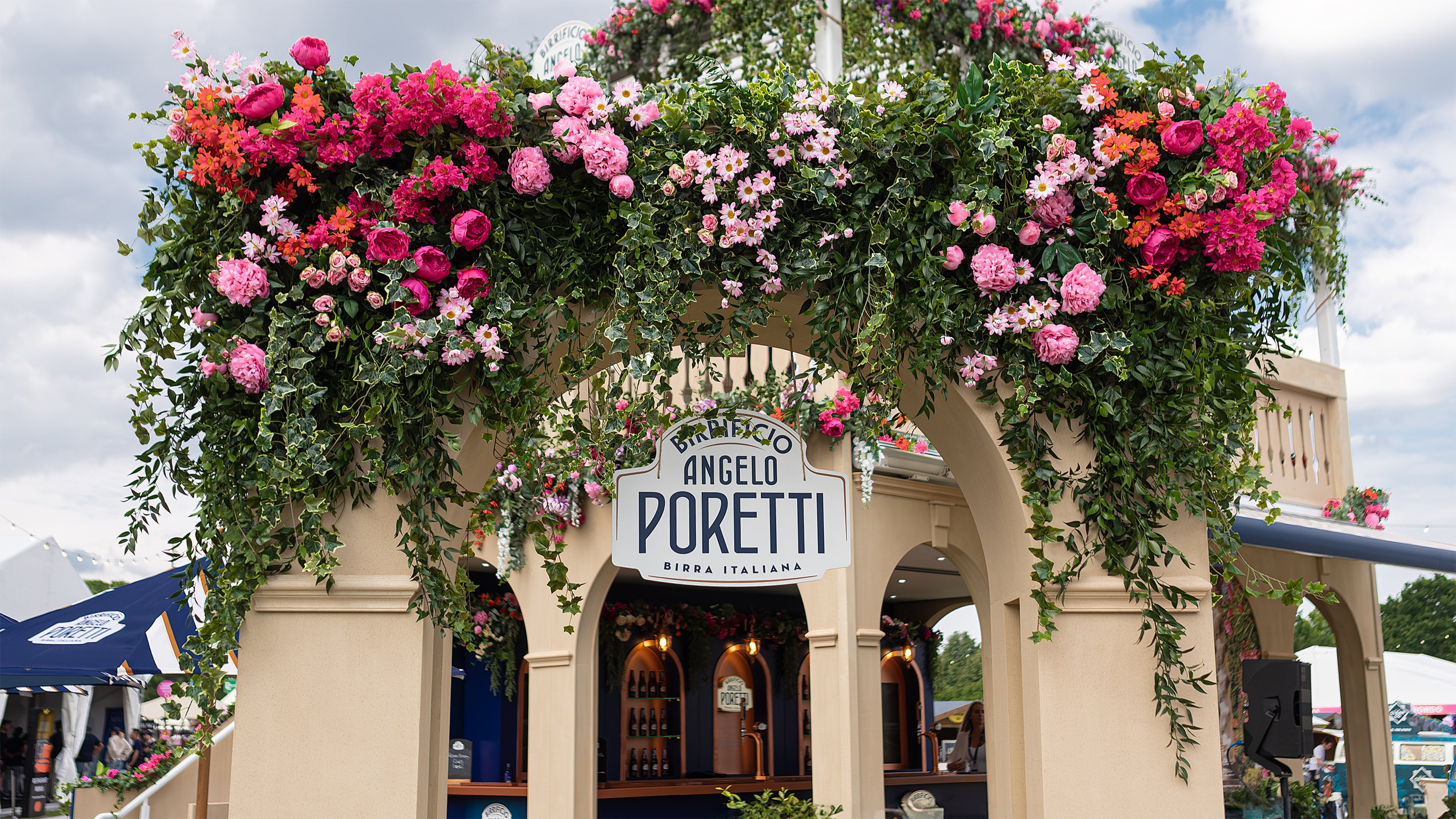 This is a close-up of the Angelo Poretti sign at Regent’s Park Taste Festival. Above the archway, lush green ivy and vibrant flowers in shades of pink, red, and orange create a welcoming atmosphere—bespoke floral arrangements by Event Florist Amaranté London.