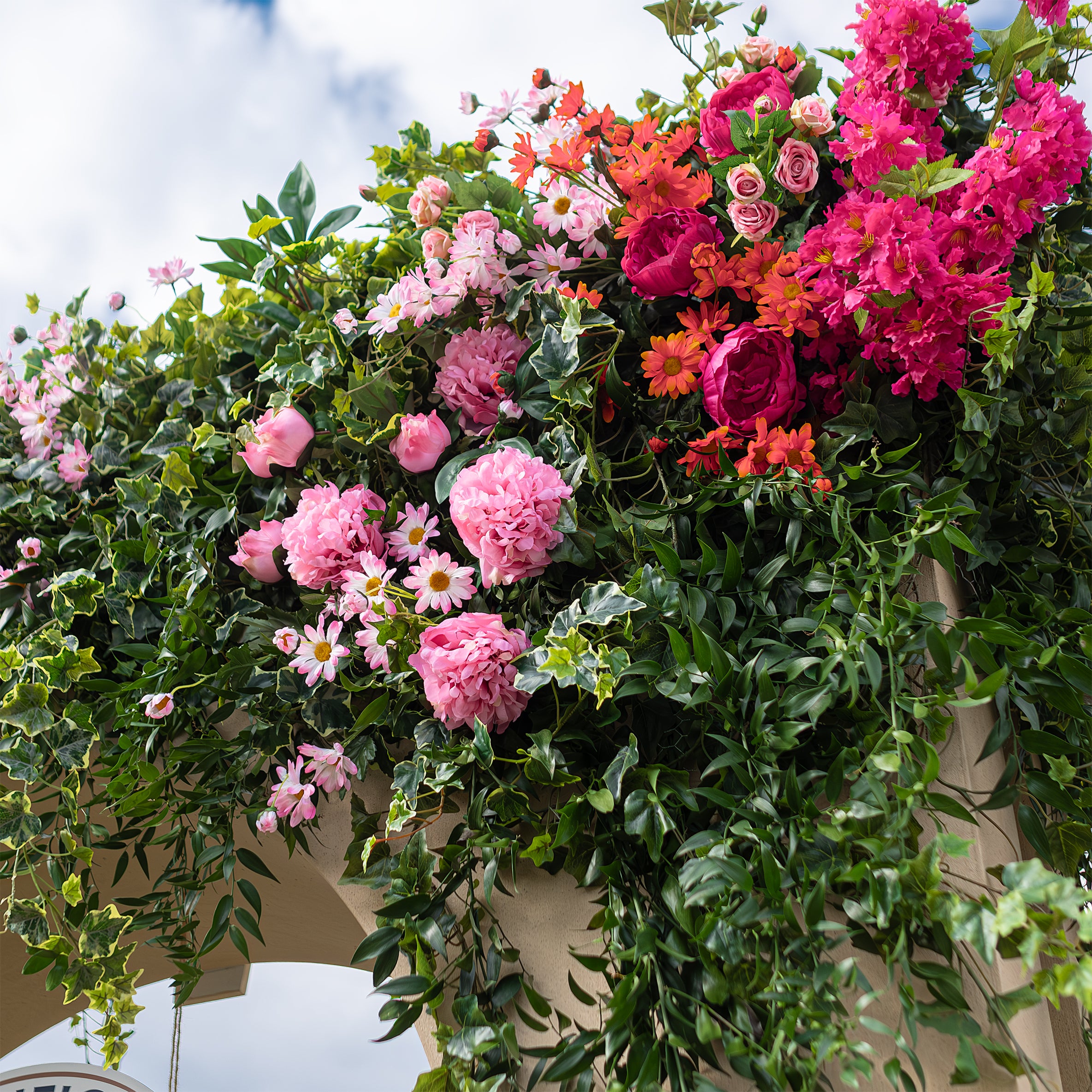 In this photo, the staircase of the Angelo Poretti event stand at Regent’s Park Taste Festival is decorated with trailing ivy and vibrant floral arrangements in pink, orange, and white by Florist Amaranté London. The floral design beautifully complements the beige stucco structure, enhancing the Mediterranean feel.