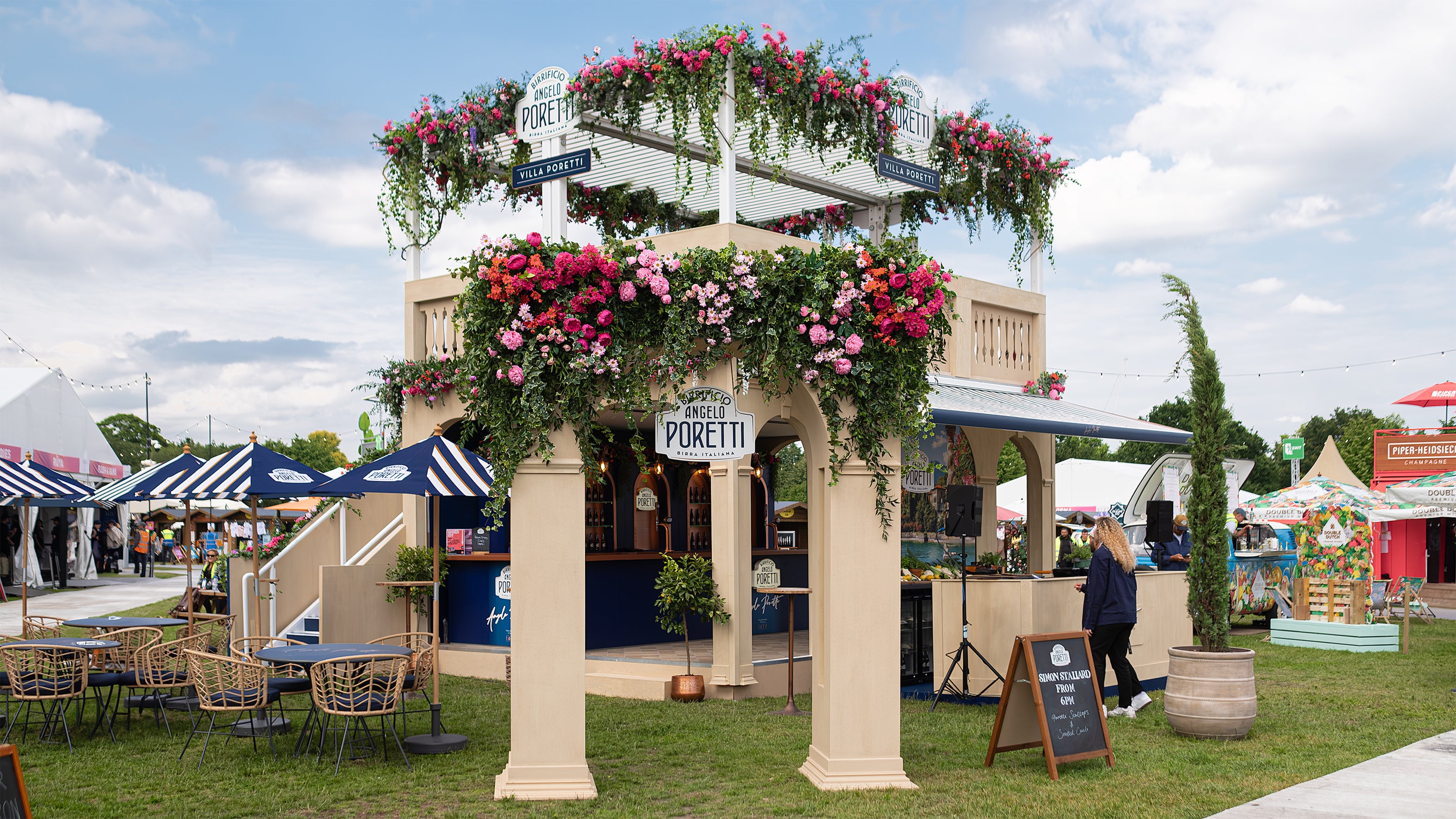 The Angelo Poretti beer stand at Regent’s Park Taste Festival Event, decorated with floral arrangements designed by Amaranté London. The archway is adorned with vibrant pink, red, and orange flowers and lush green foliage. Blue awnings and wicker chairs enhance the Mediterranean ambience.