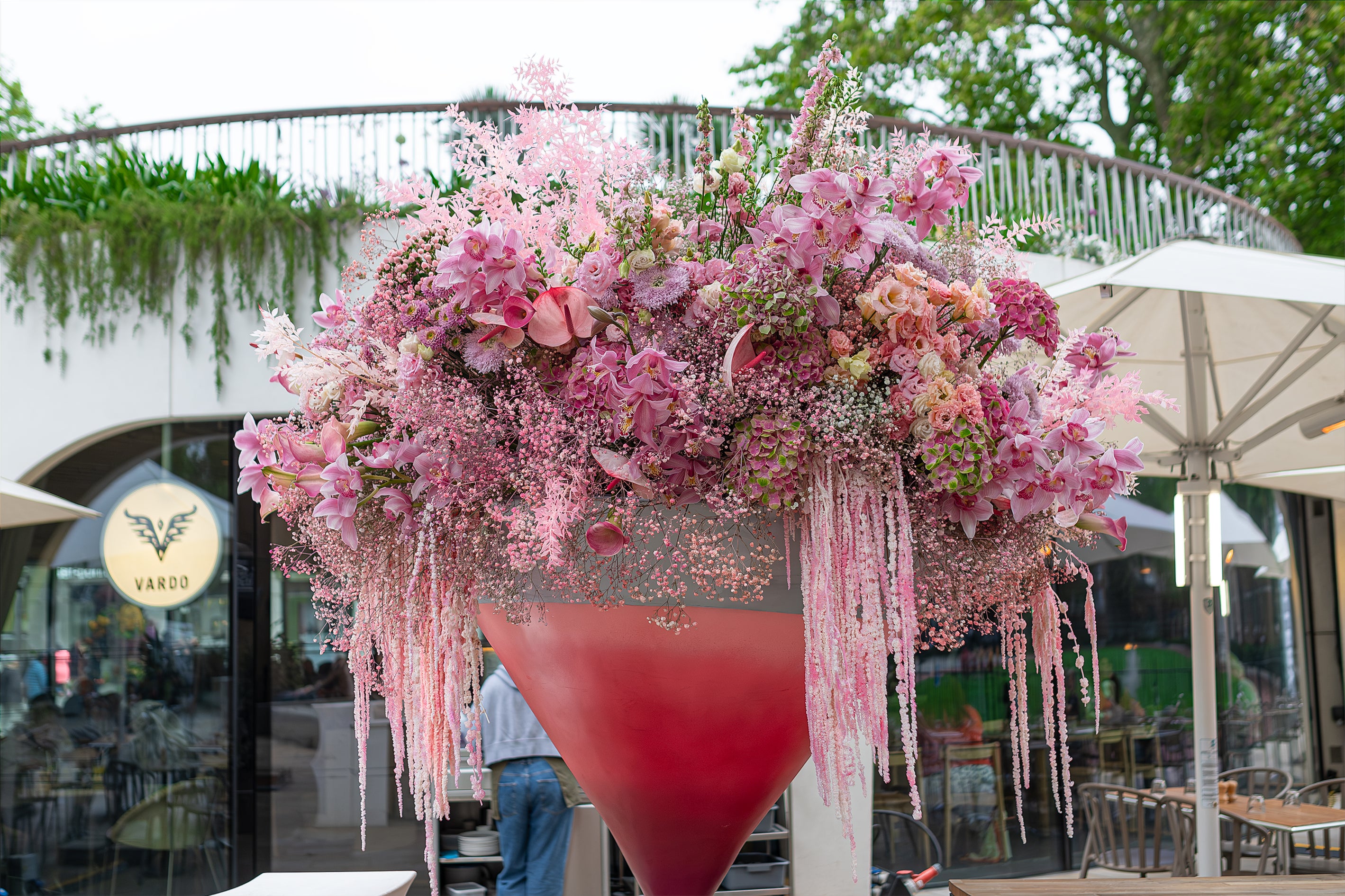 This is a close-up view of a stunning floral display in Vardo, London. Pink orchids, delicate baby's breath, and cascading blooms overflow from a giant cocktail glass also containing vibrant flowers and foliage.