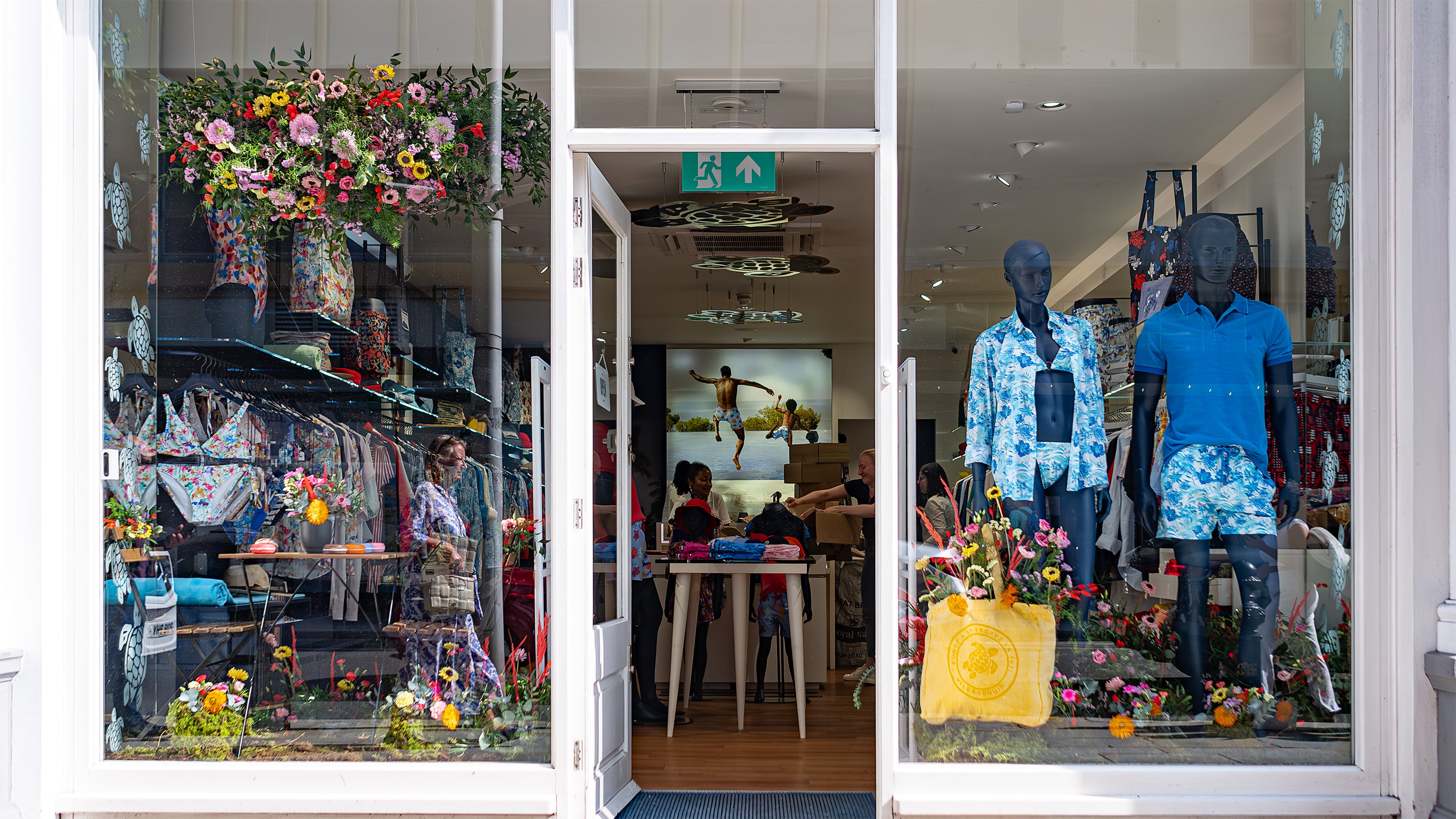 In this image, the Vilebrequin storefront is adorned with an exquisite bespoke floral arrangement designed, created and installed by event florist Amaranté London for Chelsea in Bloom. The display features a hanging floral installation and a variety of colourful flowers, enhancing the luxury swimwear boutique.