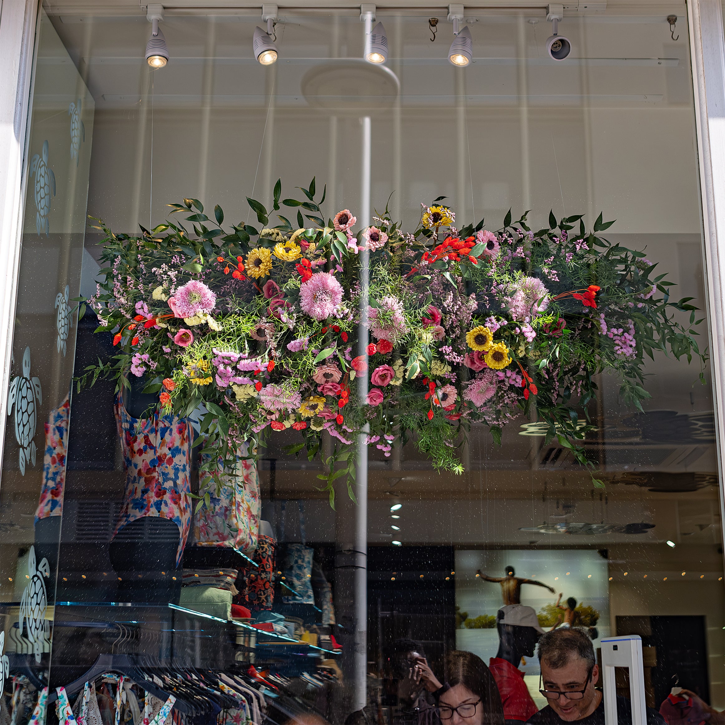 A lush hanging floral installation by Amaranté London, suspended in the Vilebrequin store window for Chelsea in Bloom, featuring a mix of pink, yellow, and red flowers, creating a whimsical and elegant display.