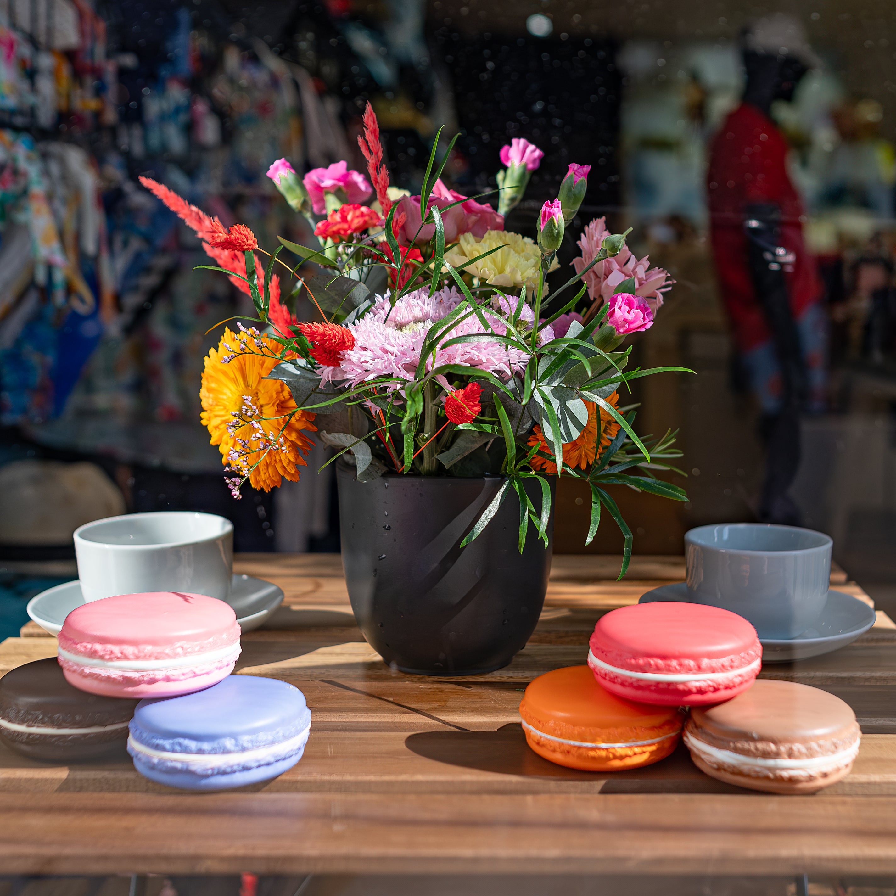 A vibrant bouquet of colourful fresh flowers in an elegant black vase placed on a table with colourful macarons enhances the Vilebrequin storefront for Chelsea in Bloom - Amaranté London.