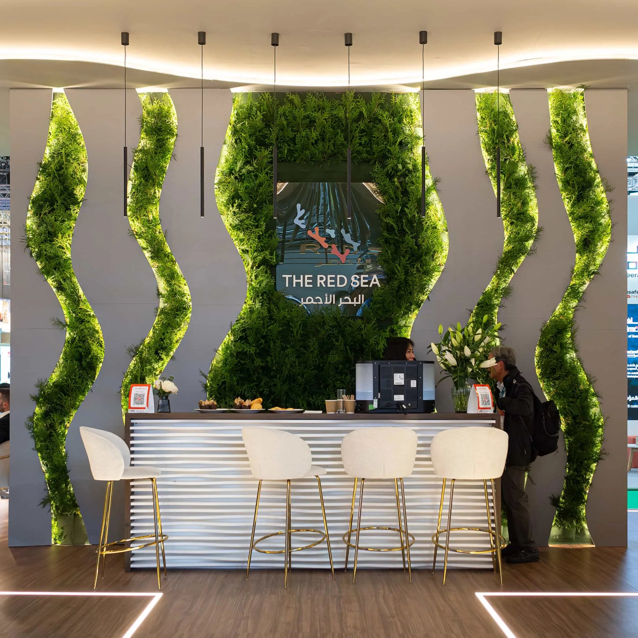 A plush green wall with a wavy design behind a bar area creates a dynamic, inviting space with integrated seating for guests to enjoy a coffee or a drink at WTM London—designed by Amaranté London.
