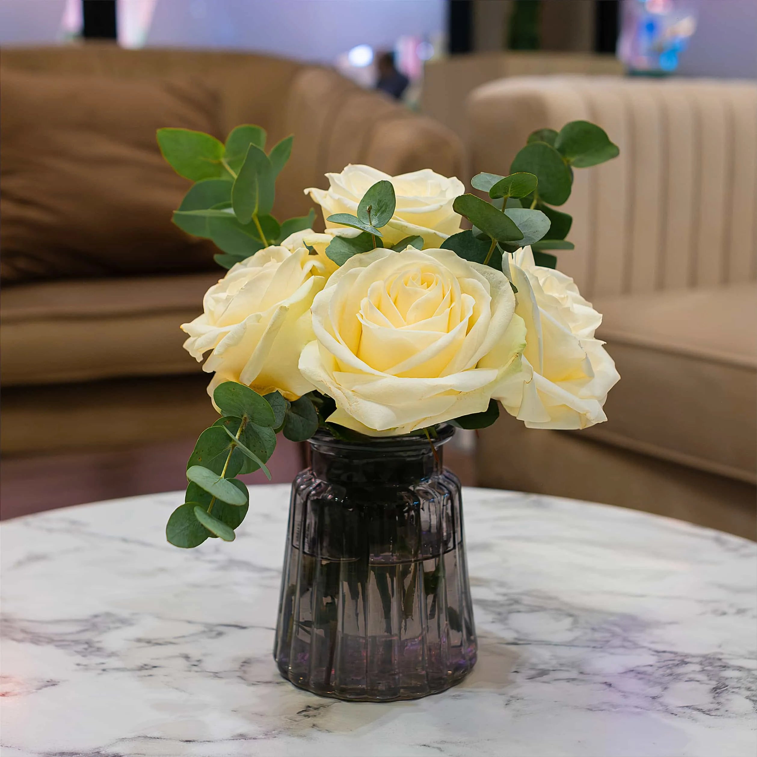This is a close-up view of a bouquet of delicate yellow roses in a dark glass vase gracing a white marble table at WTM in London.